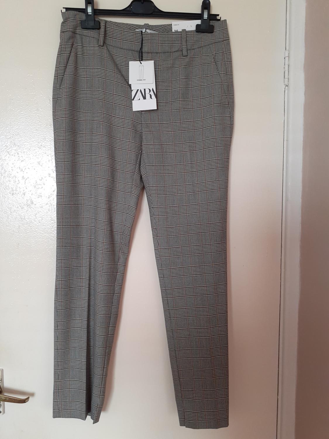 Grey checked trousers hanging on a black hanger with a Zara tag, over a white door.