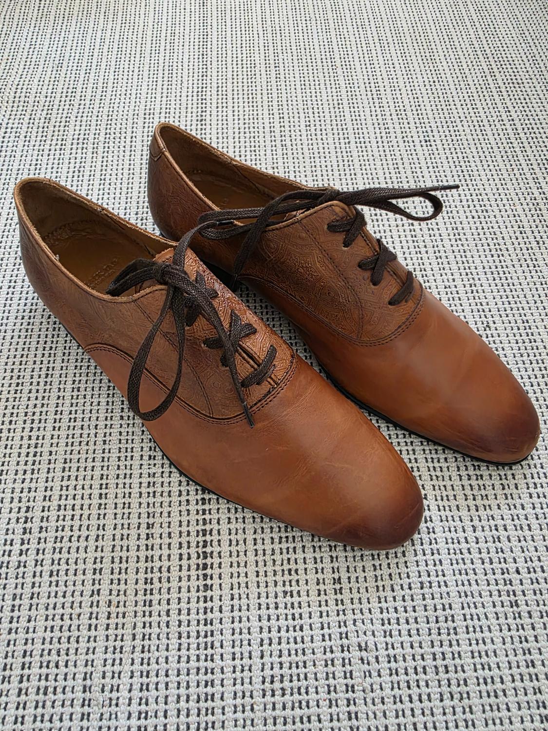 Brown leather lace-up shoes with embossed details, set on a woven surface with a black and white dash pattern.