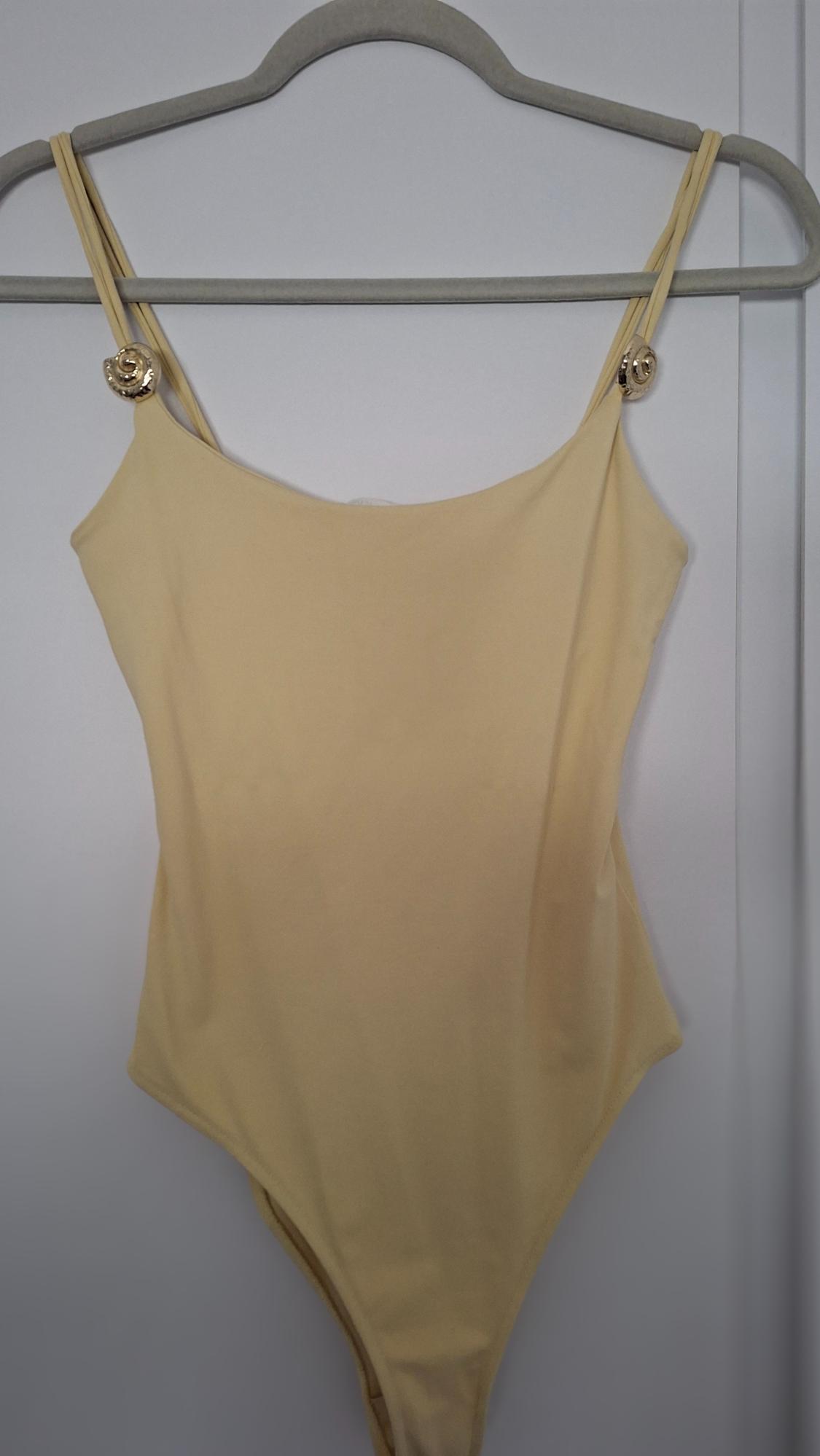 Light yellow bodysuit hanging on a grey hanger, with thin straps and gold detailing on the shoulders, against a white background.