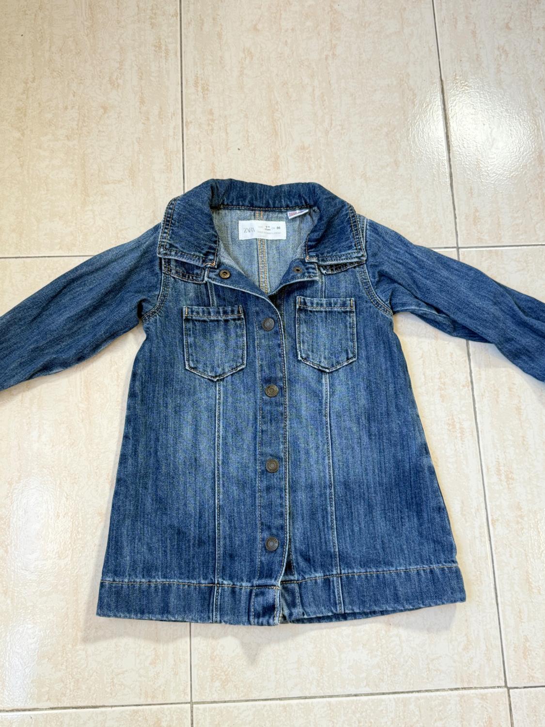 Blue denim jacket for children with front buttons and pockets, on a light tiled floor. Visible label on the collar.