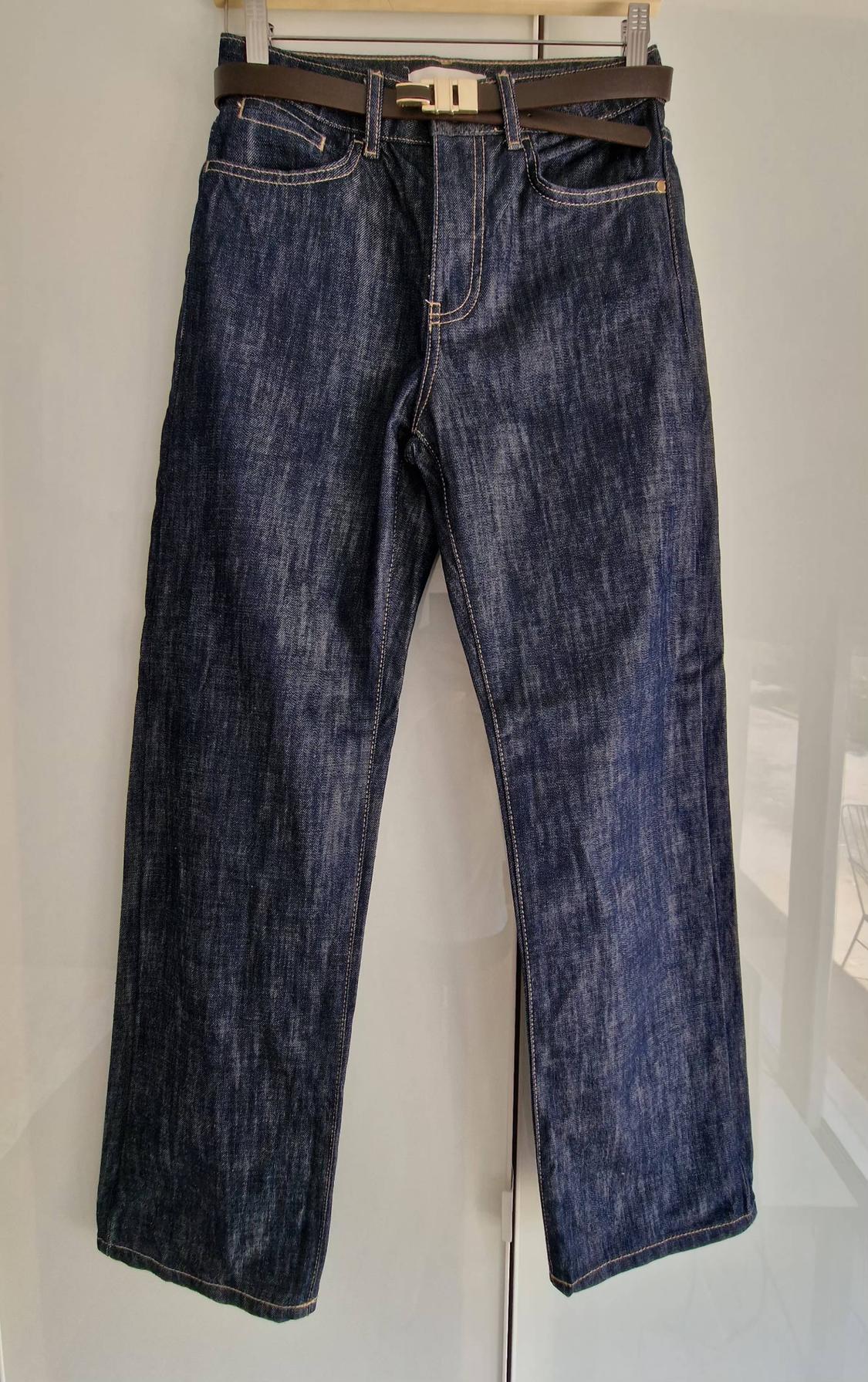 Dark blue denim jeans with light stitching and a brown belt, hanging on a hanger against a light background.