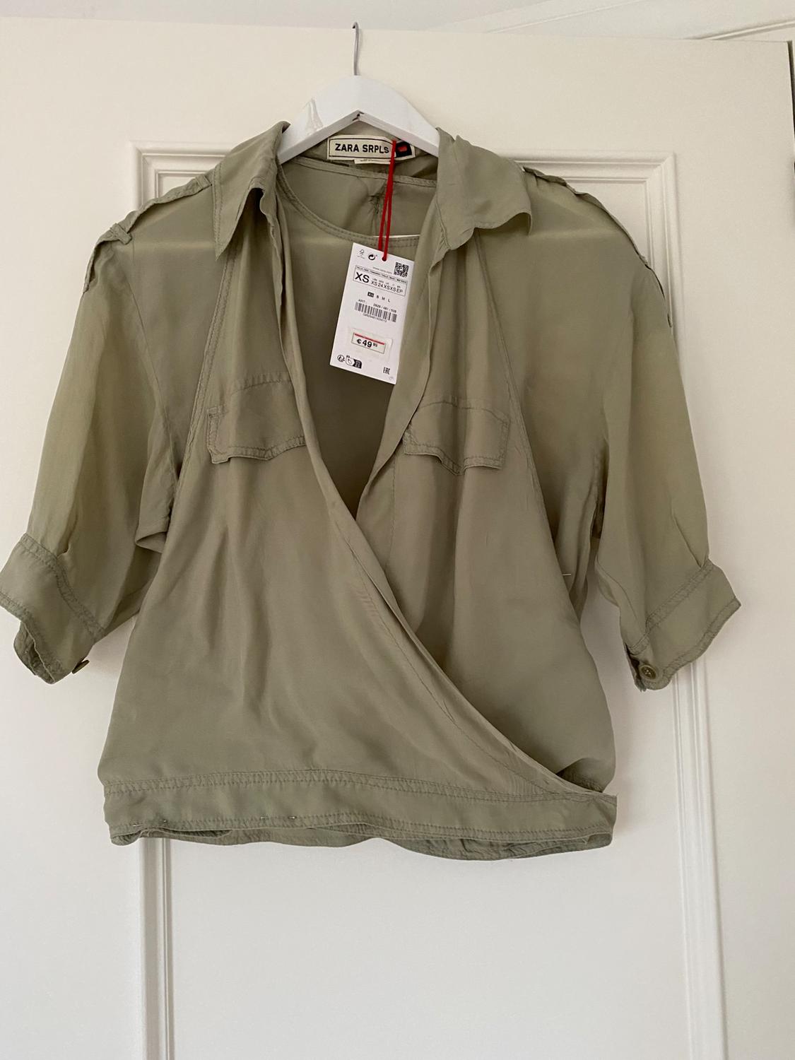 Olive green short-sleeved collared blouse, hanging on a white hanger. Visible tag with price of 49 euros. Brand ZARA SRPLS.
