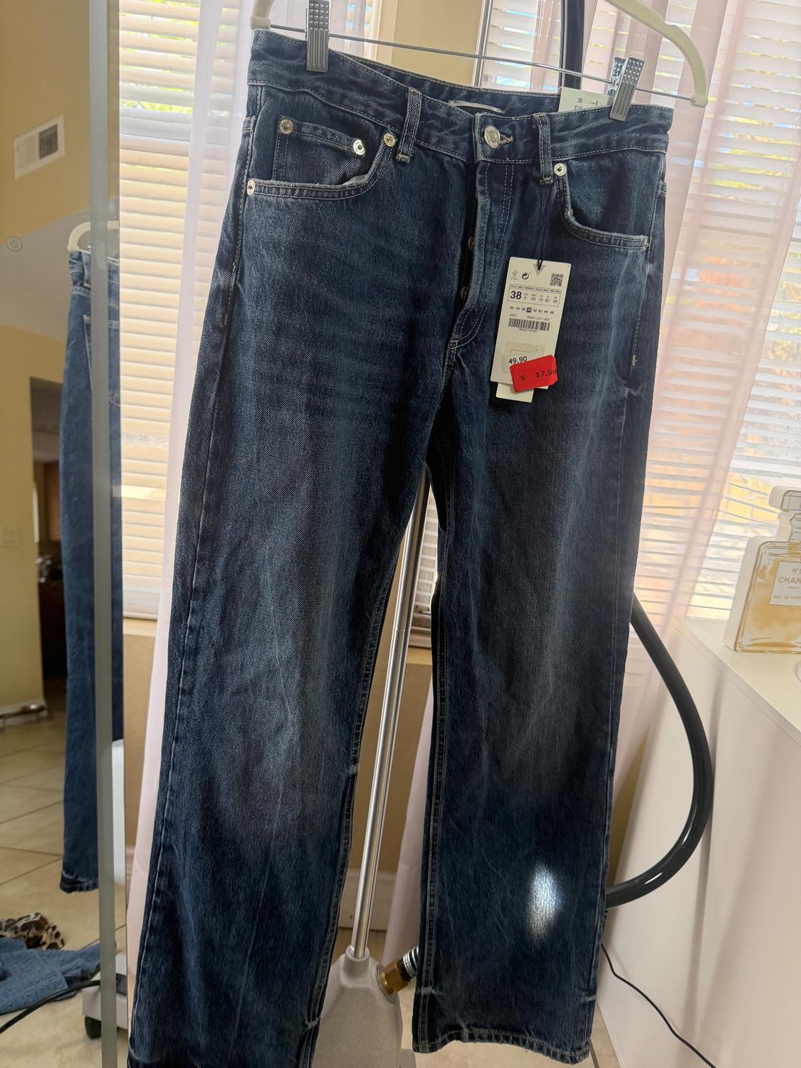 Dark blue jeans hanging on a rack, with price tags showing 49.99 and 17.99. Background with blinds and natural light.