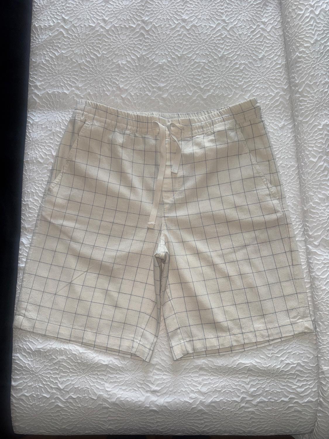 Light-coloured fabric shorts with a blue check pattern, placed on a white bedspread with a textured design.