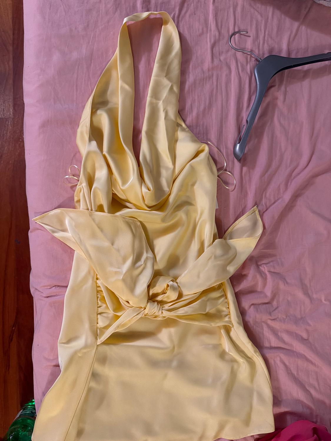 Yellow satin halter-neck dress with a front tie, laid out on a crumpled pink sheet next to a grey hanger.