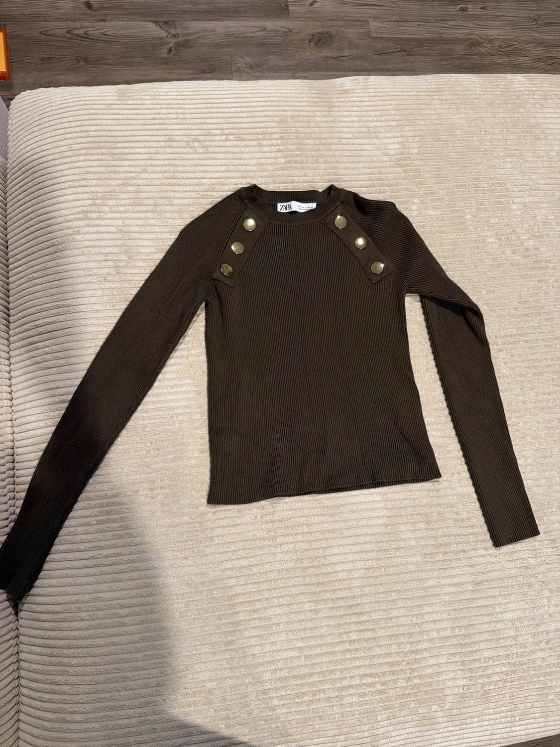 Dark brown ribbed knit long-sleeve top with gold buttons on the shoulders. ZARA label visible against a textured beige background.