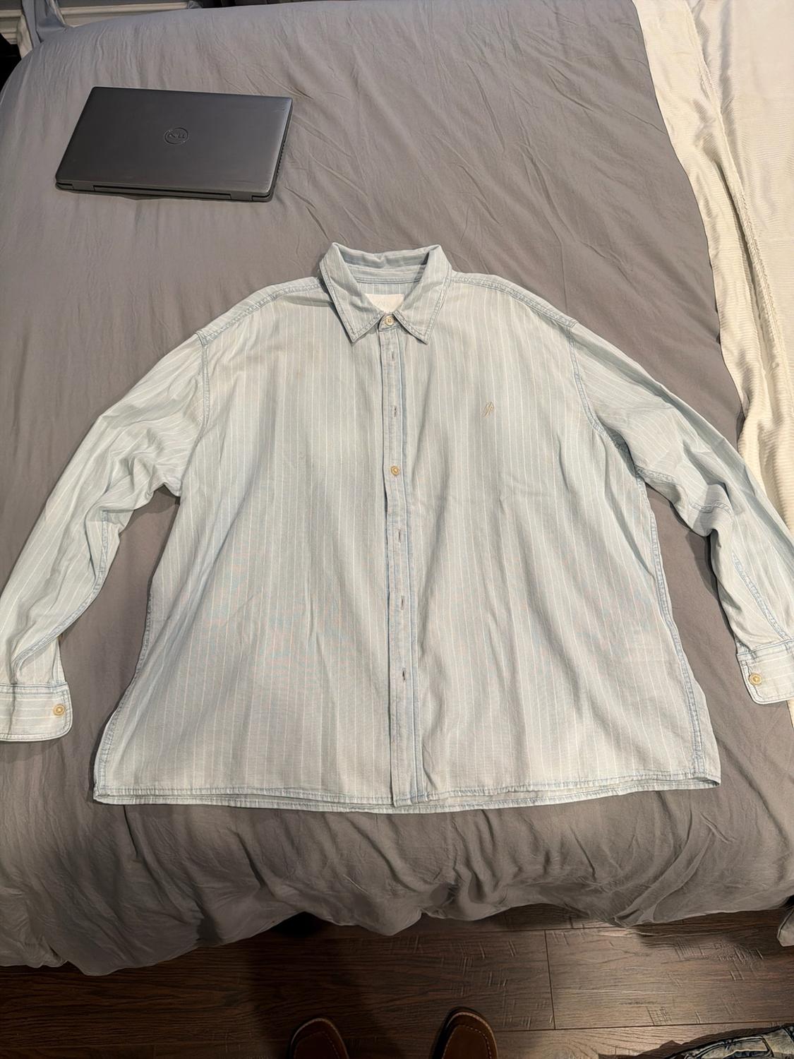 Light blue long-sleeved shirt with fine stripes on a grey bed. A laptop and shoes complete this relaxed composition.