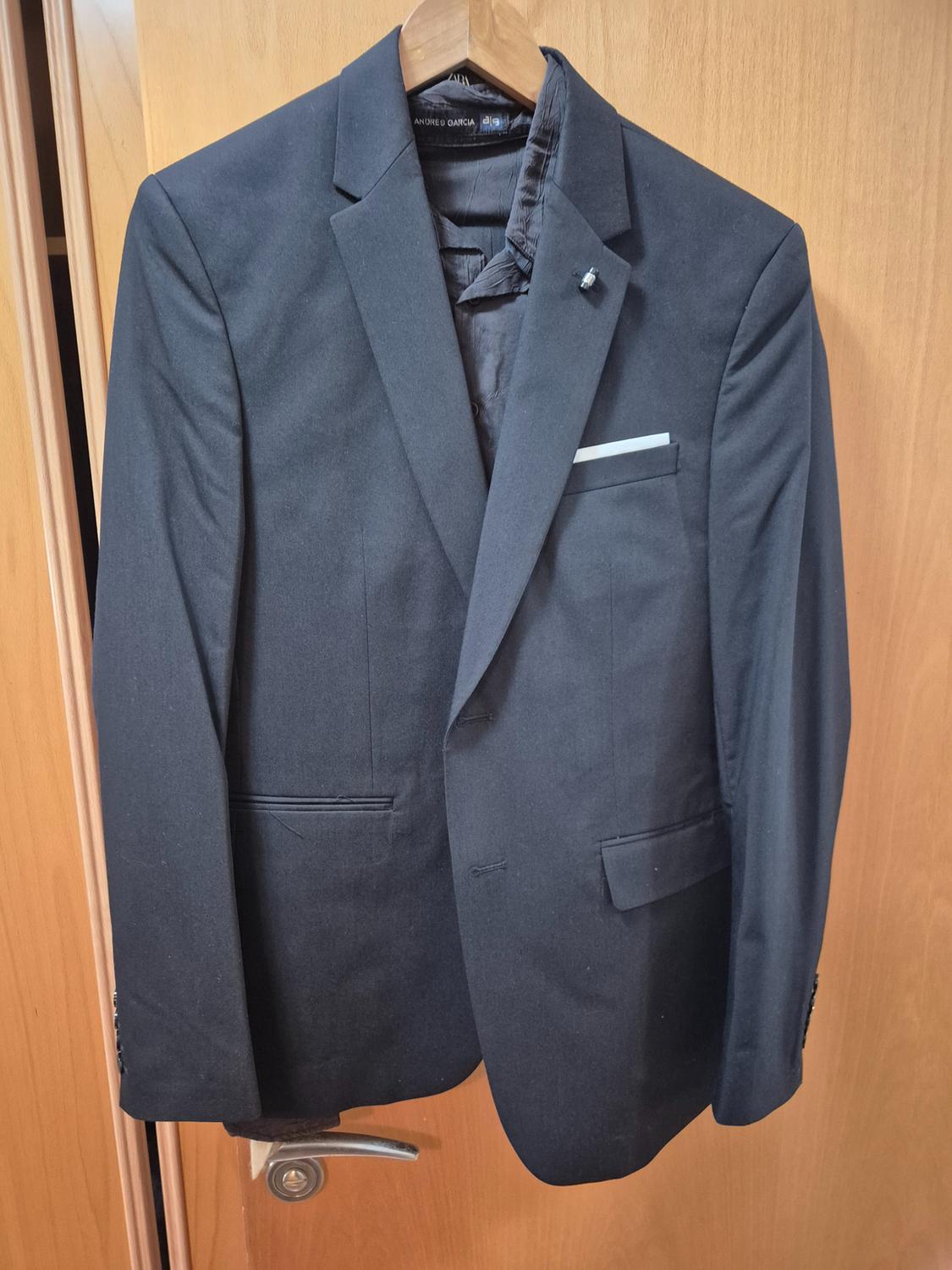 Black suit jacket hanging on a wooden hanger over a light wooden door. Pocket with white handkerchief visible.