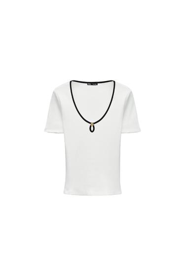 Front view of a white t-shirt with black trim on the collar and brooch detail in the centre.