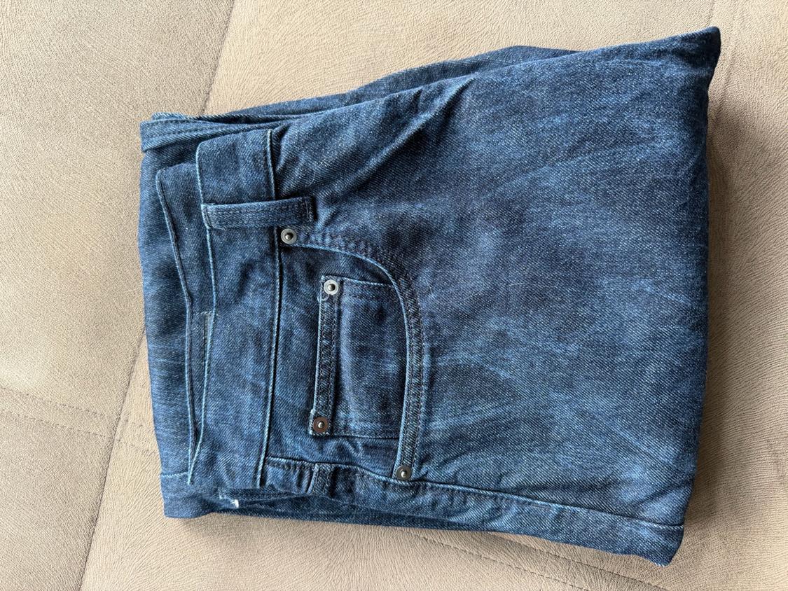 Dark blue jeans folded on a beige fabric surface, highlighting the front pocket with rivets and stitching.