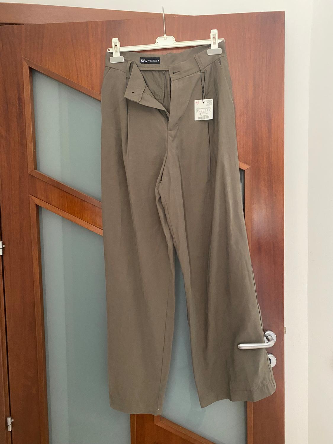 Khaki wide-leg trousers hanging on a wooden door. Visible tag with text: "Zara Woman".