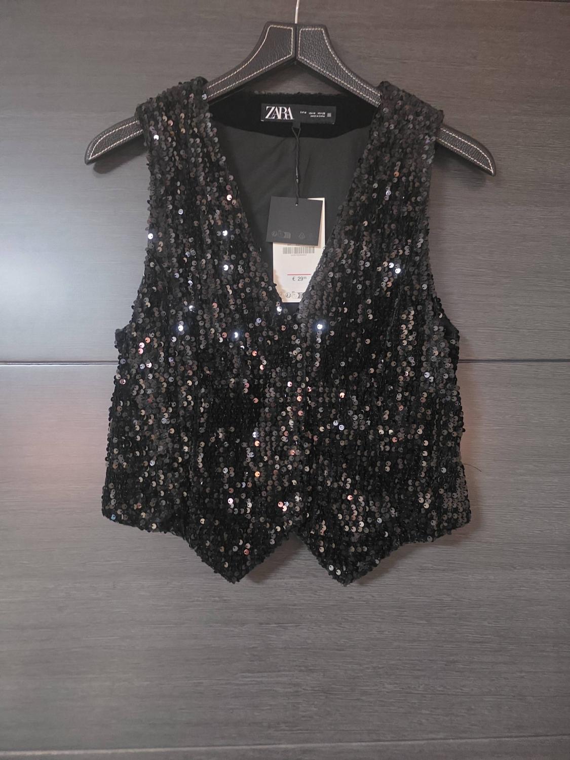 Black sequinned waistcoat hanging on a coat hanger over a dark wooden door. Visible label with the text ZARA.
