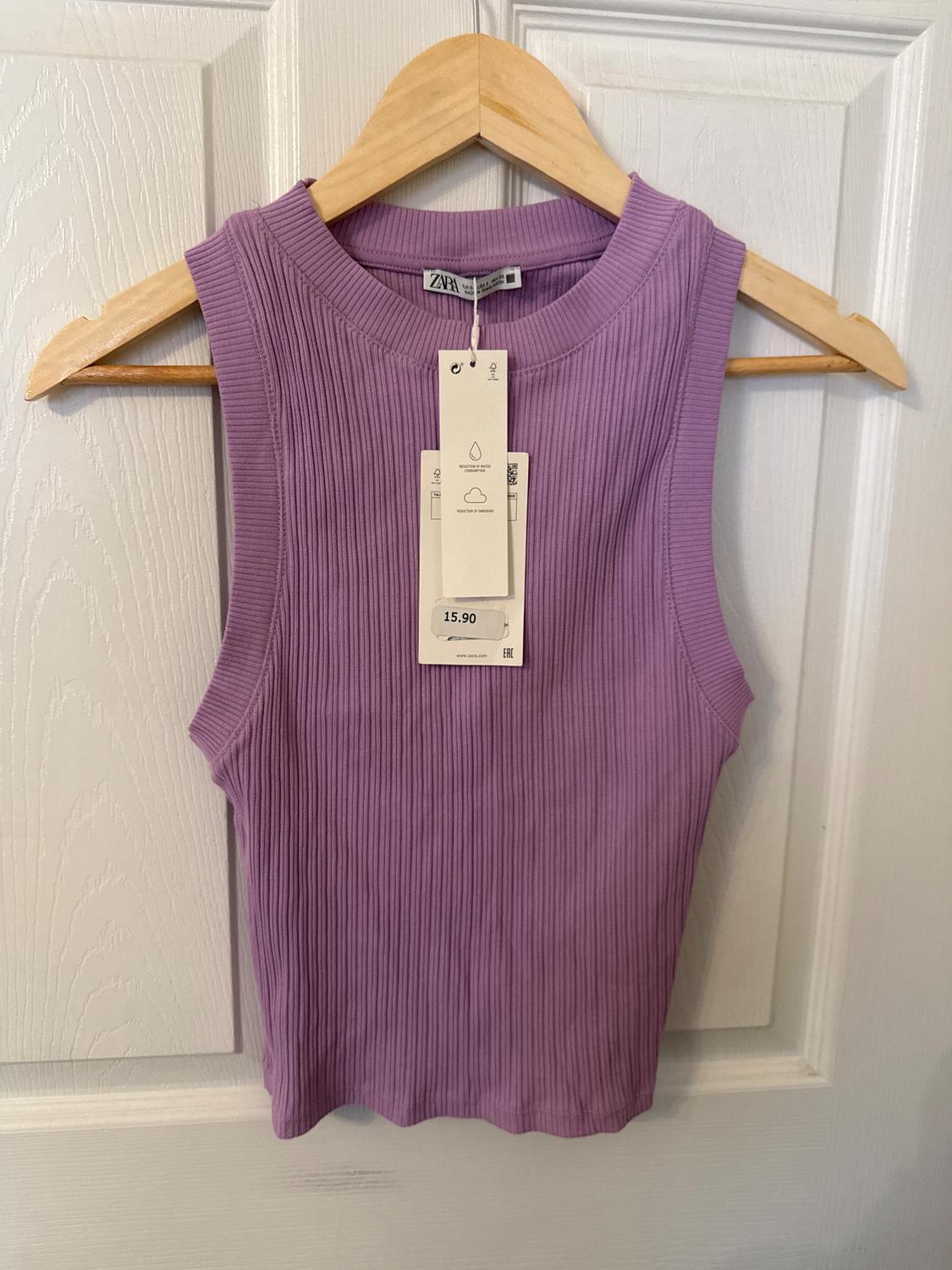 Lilac ribbed sleeveless top hanging on a wooden hanger. Visible tag with a price of 15.90. White door background.