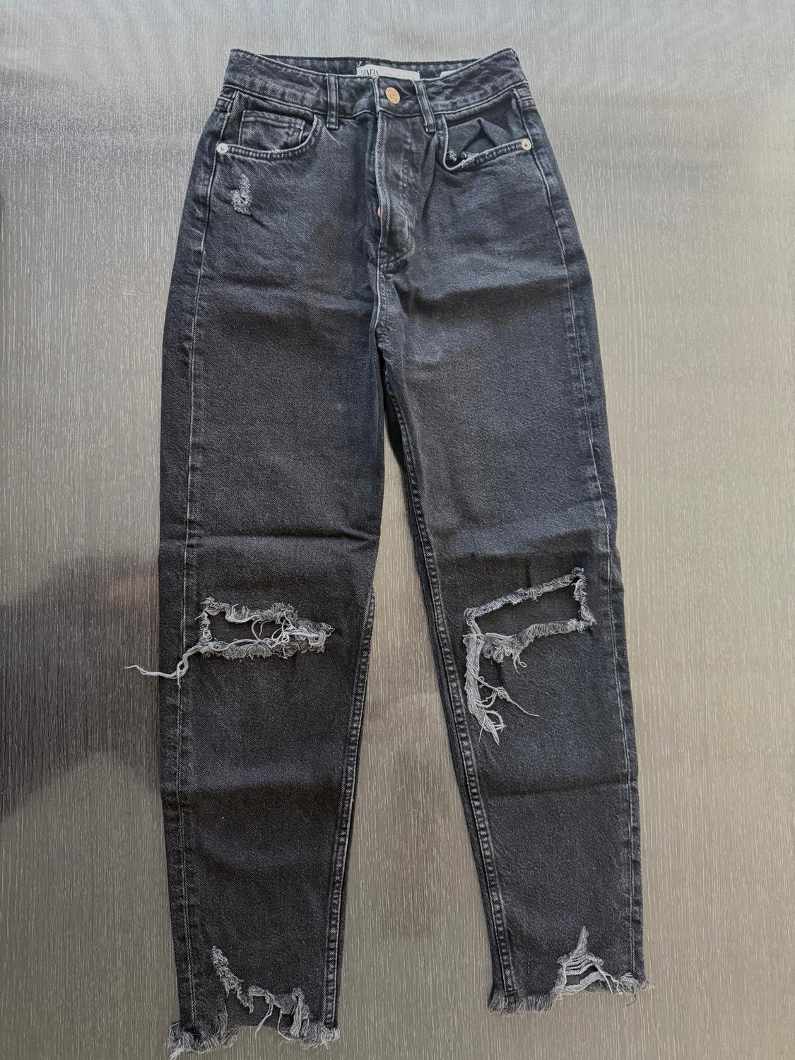 Distressed black jeans with rips at the knees and ankles, on a textured gray background.