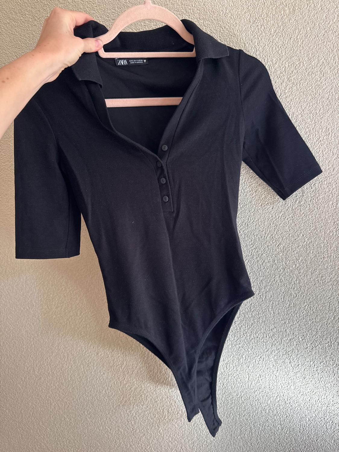 Hand holding a black short-sleeved bodysuit with a collar and buttons on a pink hanger against a light-coloured wall. Visible text: ZARA.