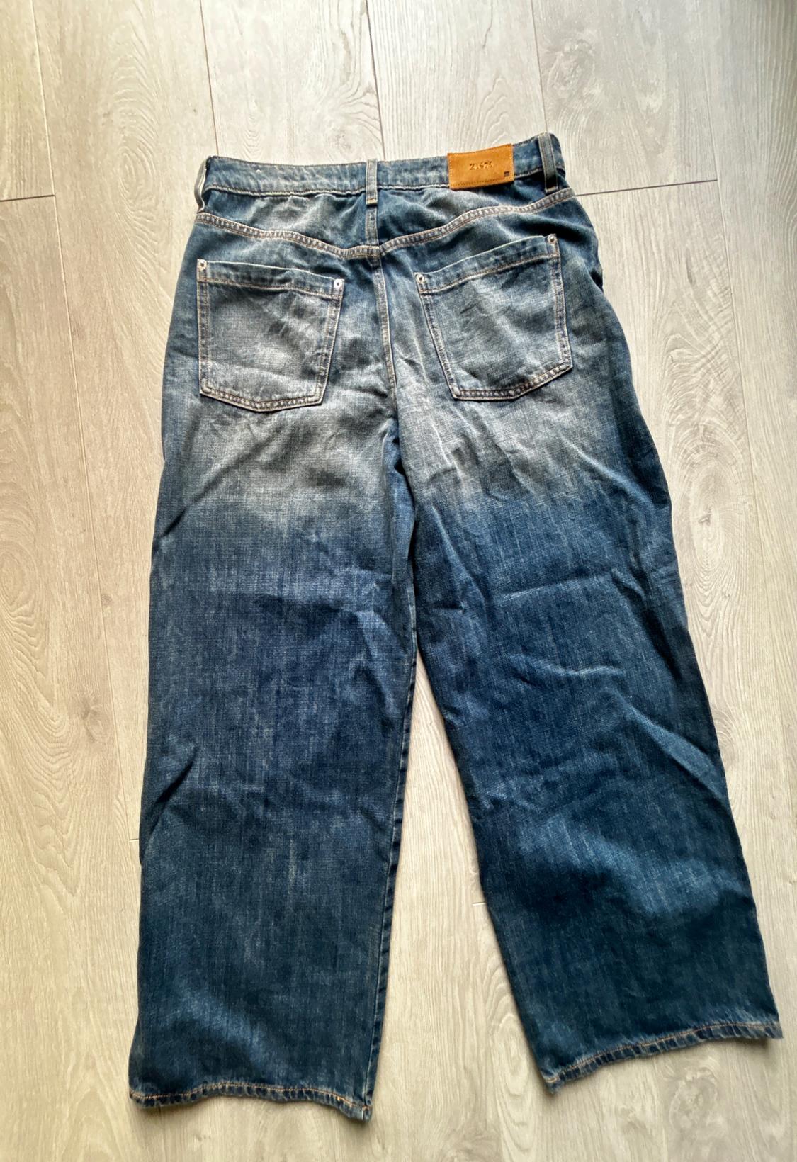 Blue distressed denim jeans seen from behind, with pockets and Z1975 label, laid out on a light wooden floor.