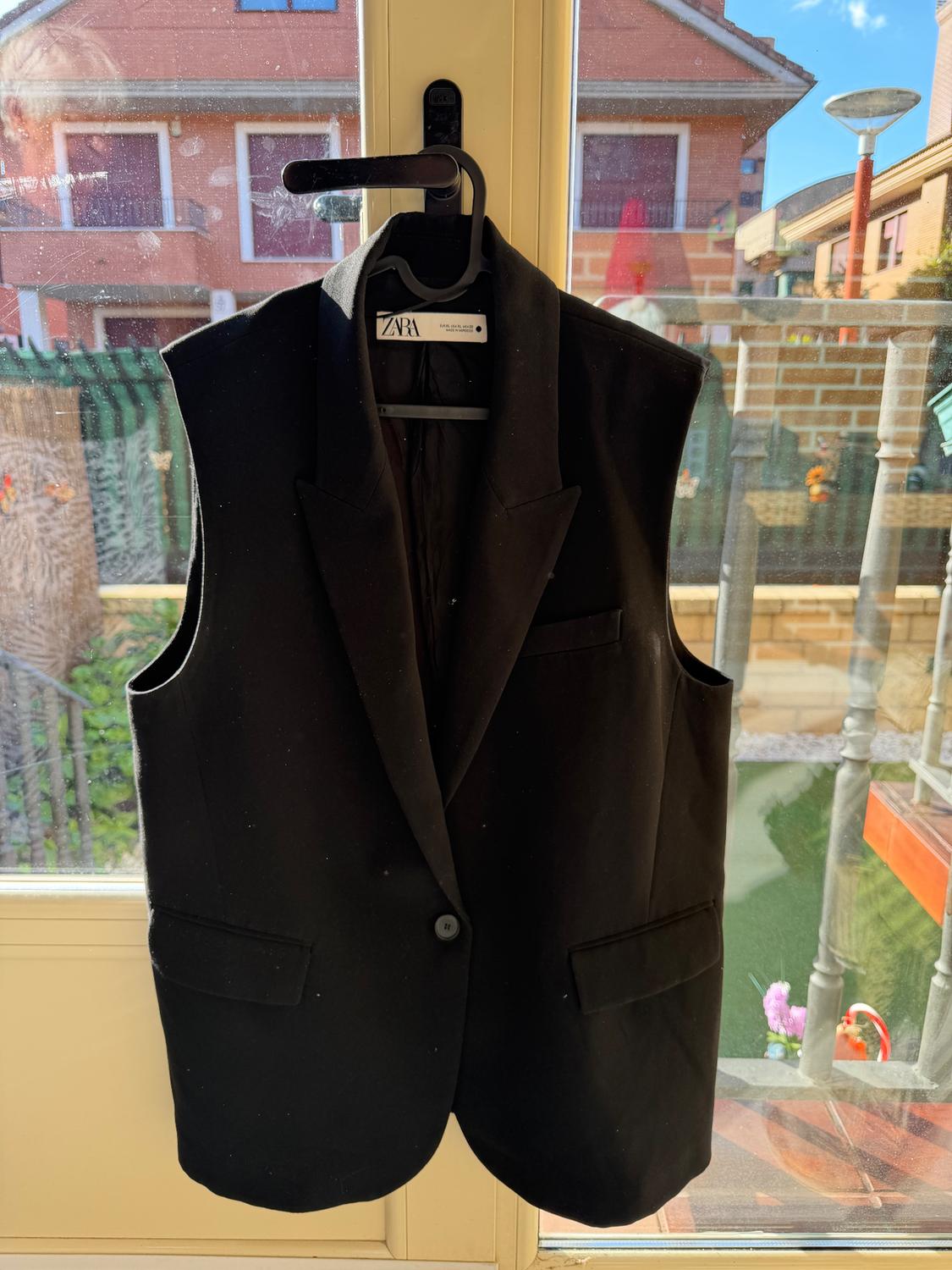 Black waistcoat hanging on a hanger in front of a window, with a visible Zara tag. Background of houses and blue sky.