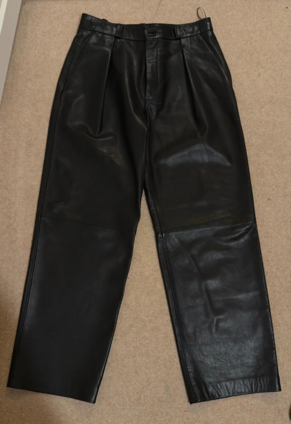 Black leather trousers with front pleats, placed on a beige rug.