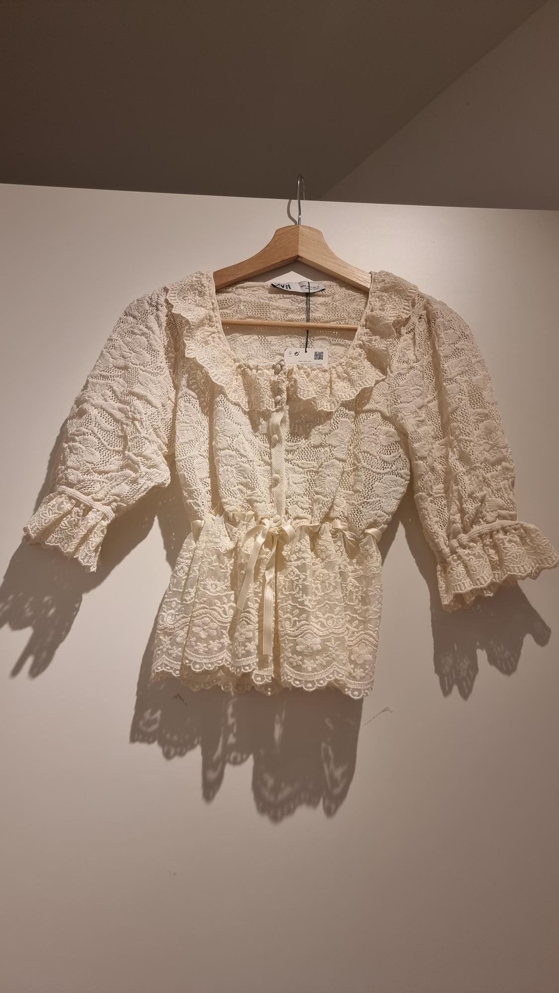 Beige lace blouse with three-quarter sleeves and a tie at the waist, hanging on a wooden hanger against a white background.