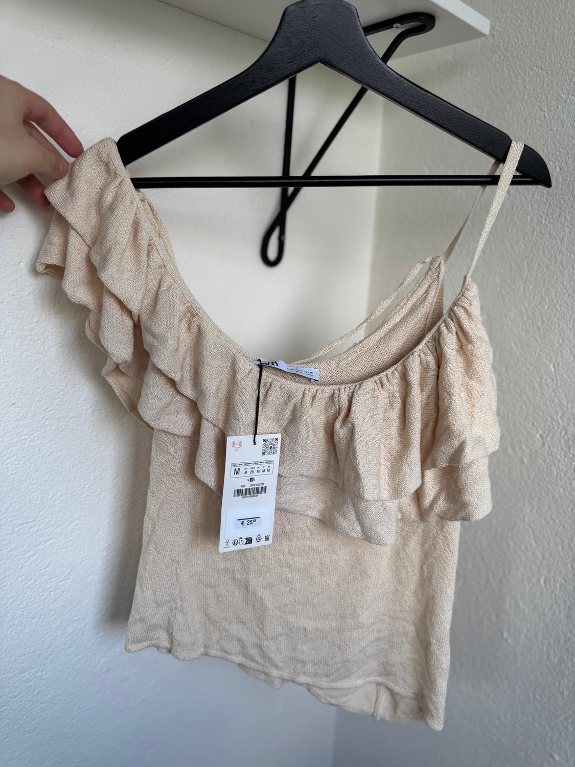 Beige ruffled top on black hanger, visible tag with price of €25. White background and hand holding the hanger.