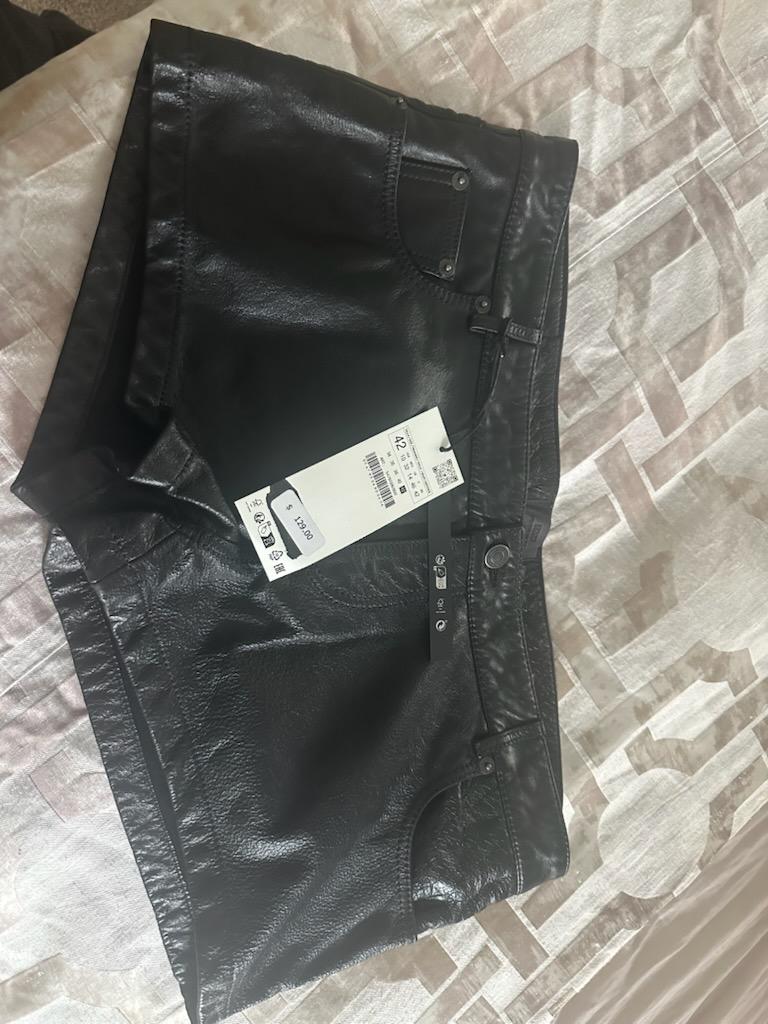 Black shiny material shorts on a bed, with a visible price tag showing 120.00.