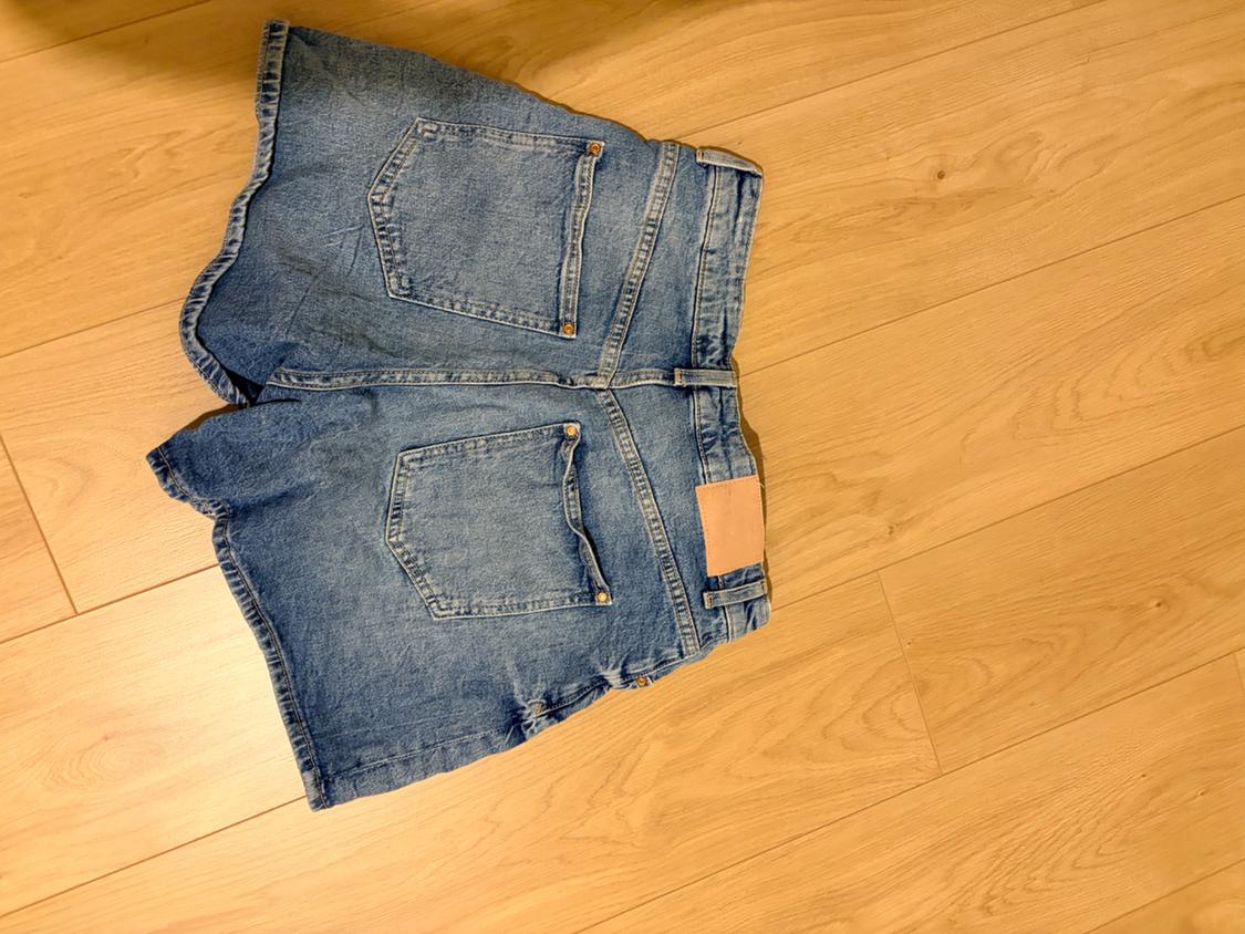 Blue denim shorts seen from behind, with pockets and a patch on the waistband, on a light wood floor.