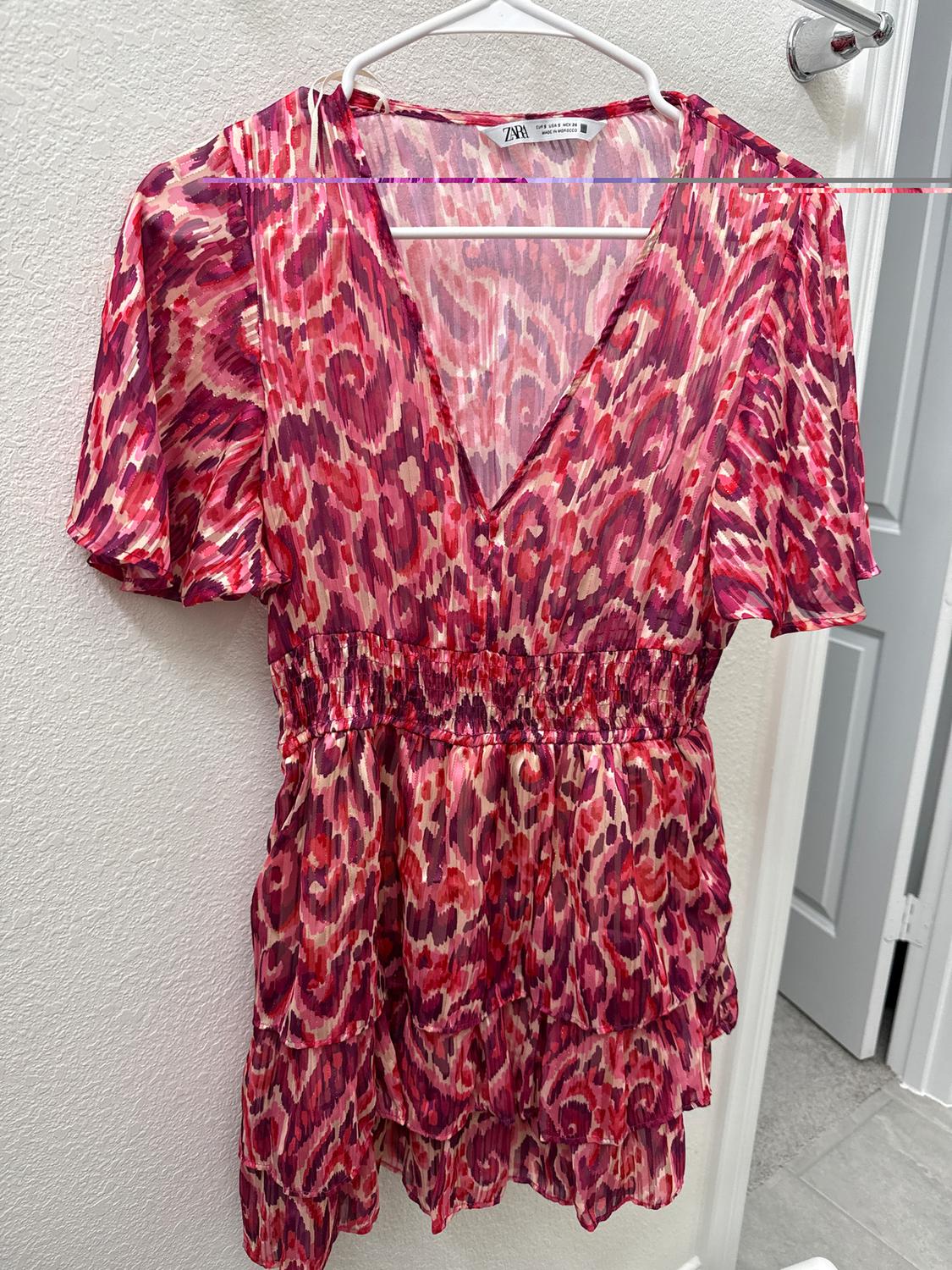 Short dress with an abstract print in pink and red tones, hanging on a white hanger against a light-colored wall.