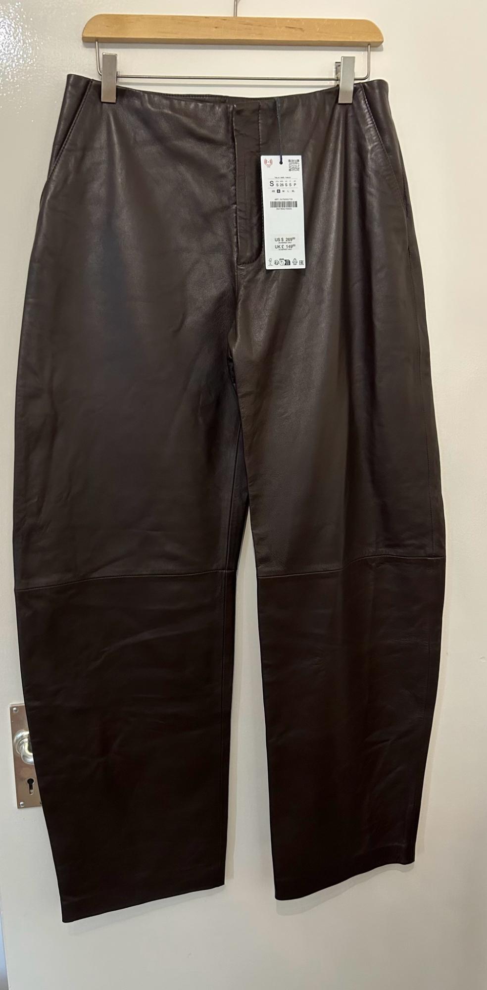 Dark brown wide-leg trousers hanging on a wooden hanger against a white door. Label with text: S 26 S S P US $ 269.00 UK £ 149.00.