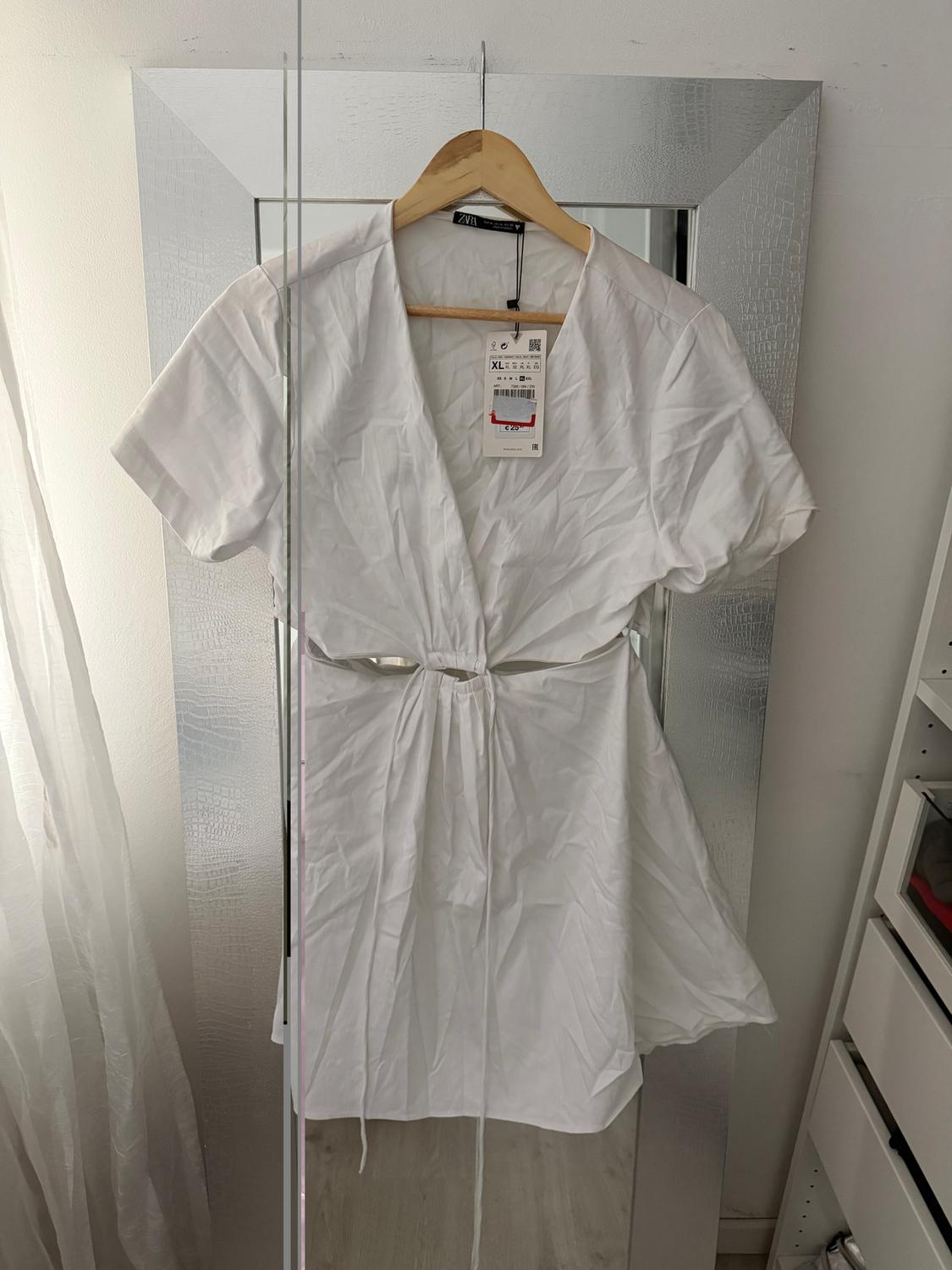 White dress hanging on a hanger in front of a mirror. Visible tag with size XL. Background with white curtain and shelving unit.