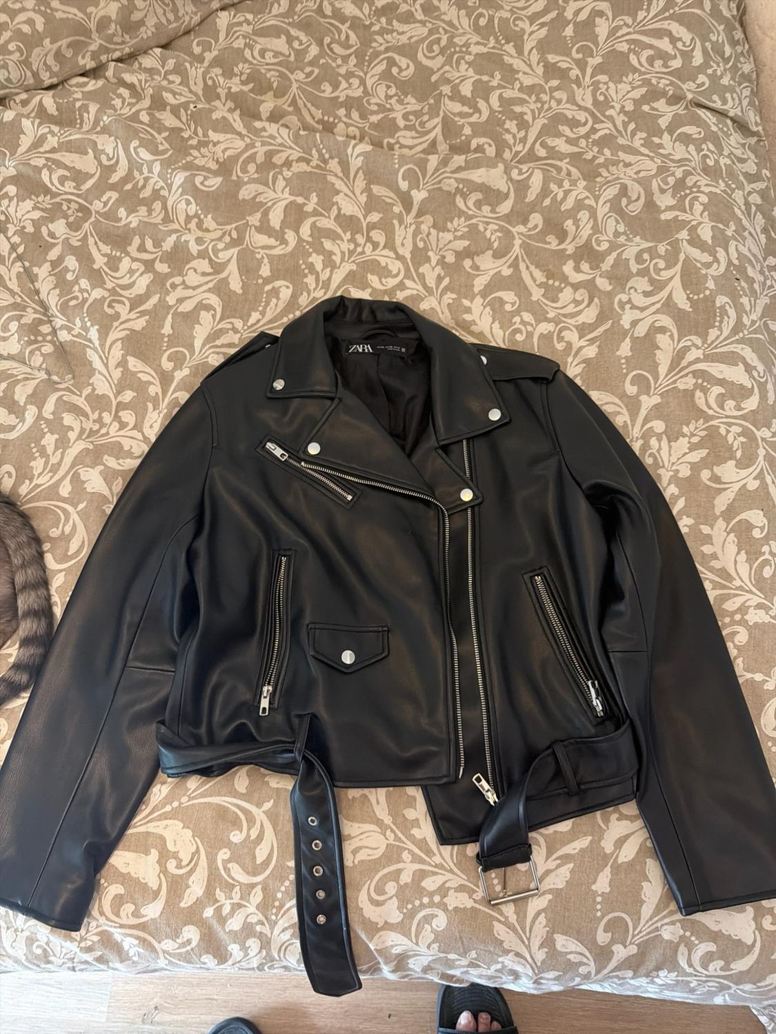 Black biker jacket with zips, a belt and a ZARA label, on a bed with a floral-print bedspread.