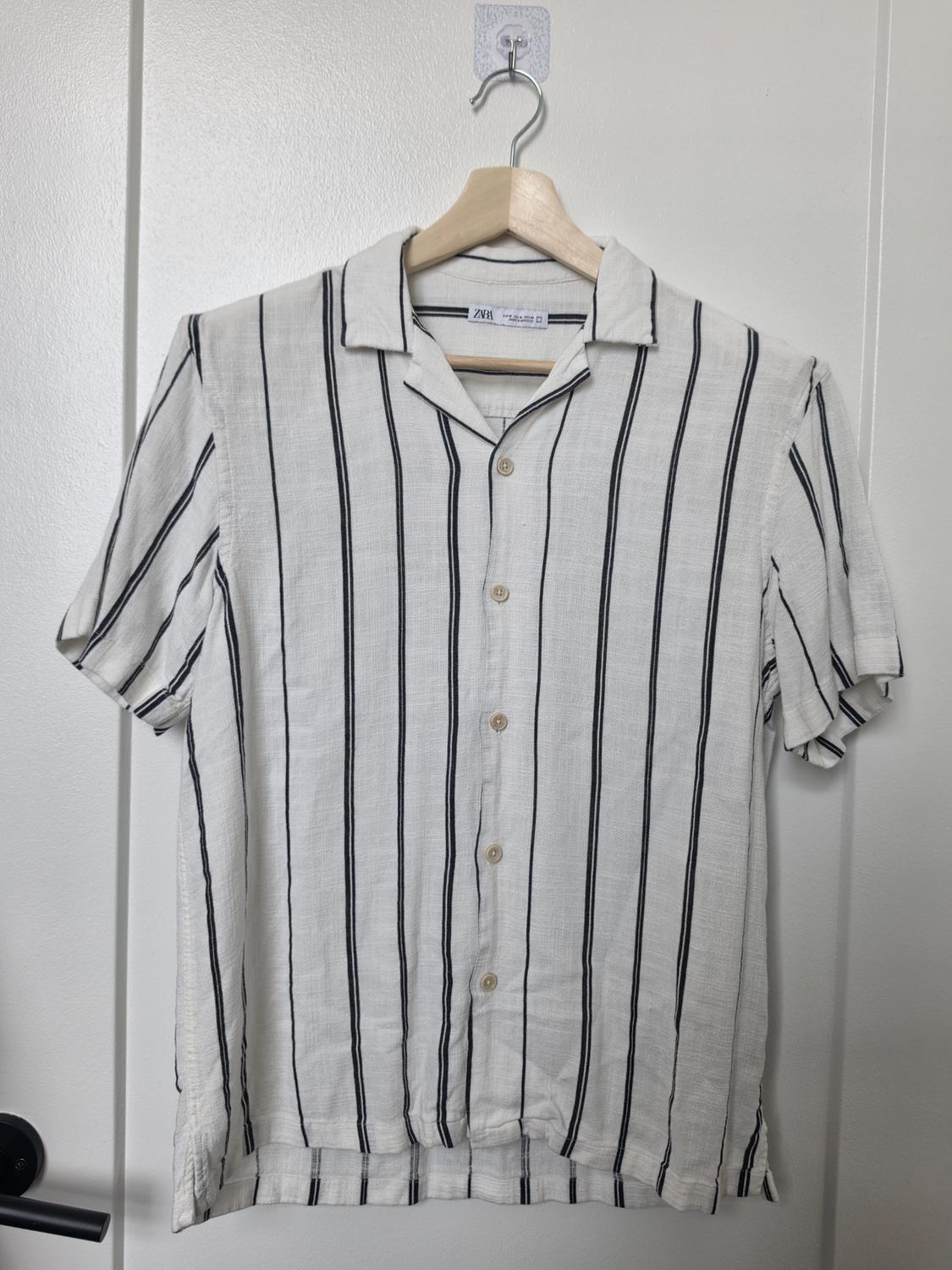 White short-sleeved shirt with black vertical stripes hanging from a wooden hanger on a white door. Text: ZARA.
