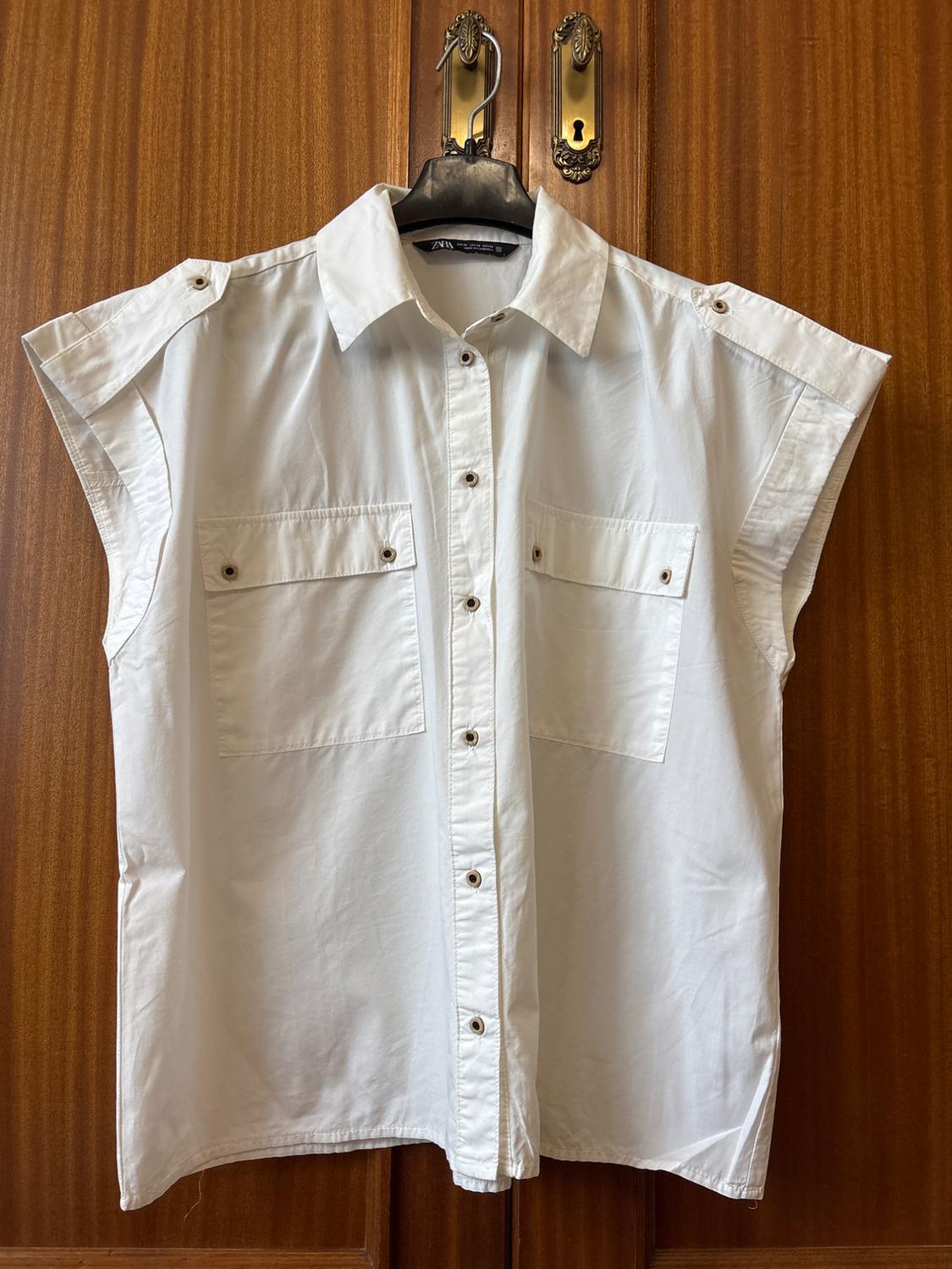 White sleeveless shirt with buttons and two front pockets, hanging on a hanger over a wooden door.