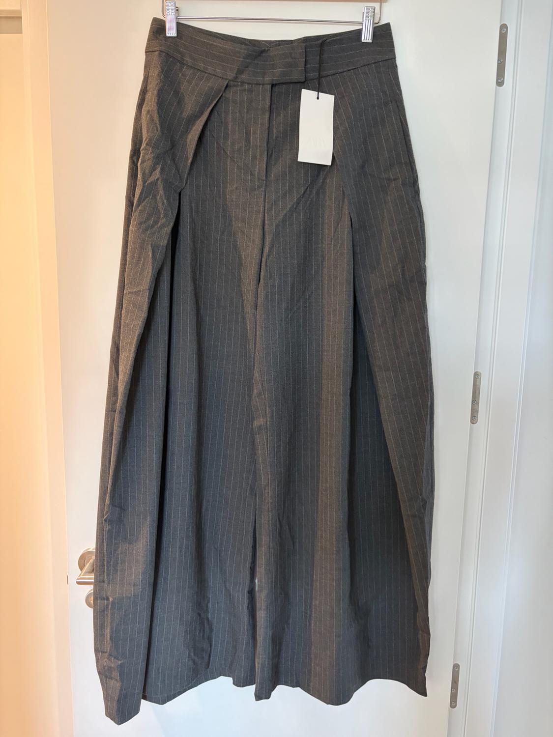 Σκούρο γκρι παντελόνι με ρίγες (pinstripe), σε φαρδιά γραμμή, κρεμασμένο σε μια κρεμάστρα πάνω από μια λευκή πόρτα. Διαθέτει μια εμφανή ετικέτα.