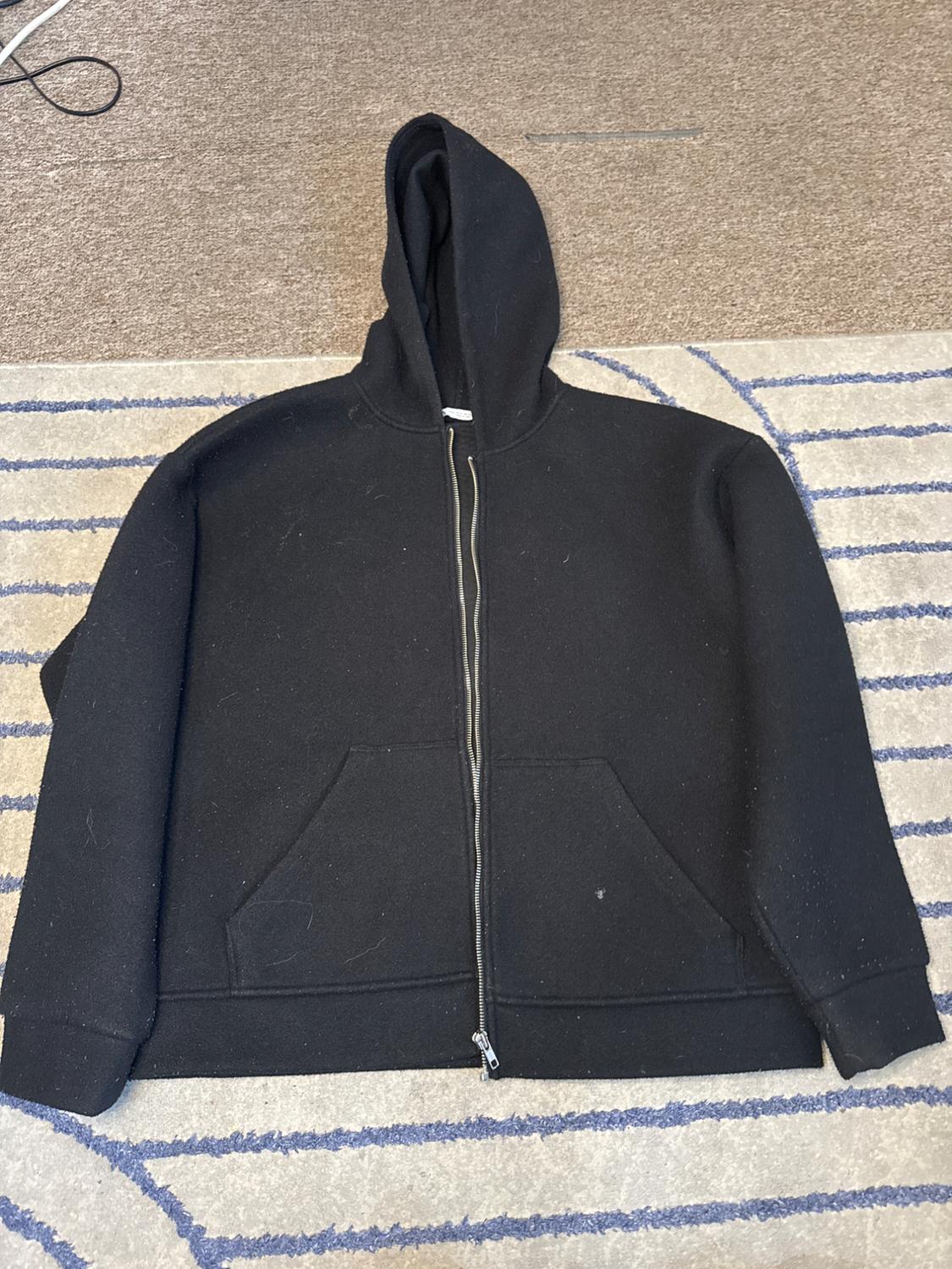 Black hoodie with a zip and pockets, on a beige rug with blue lines. Relaxed composition with a casual style.