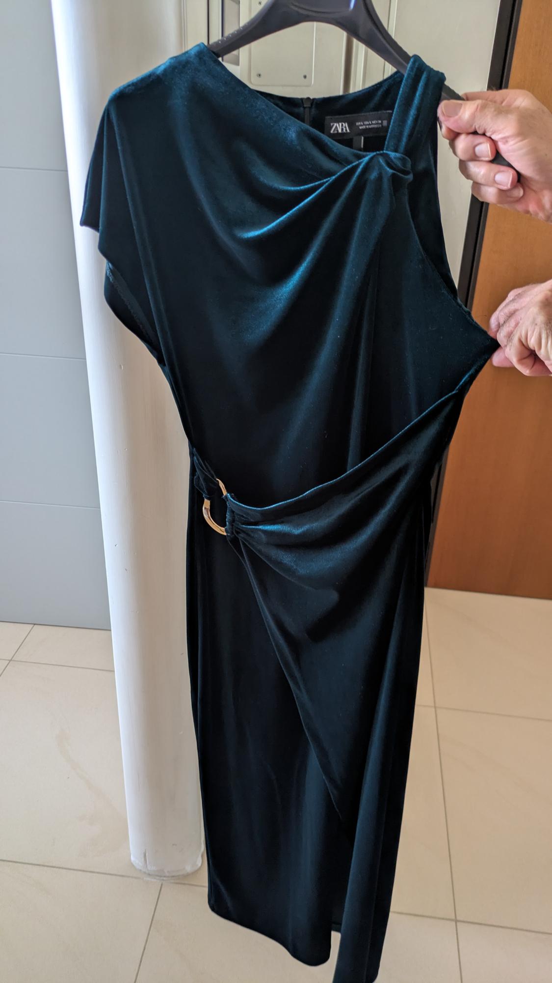 Hands hold a dark green velvet dress on a hanger. It features a draped design, gold hoop, and ZARA tag. Indoors.