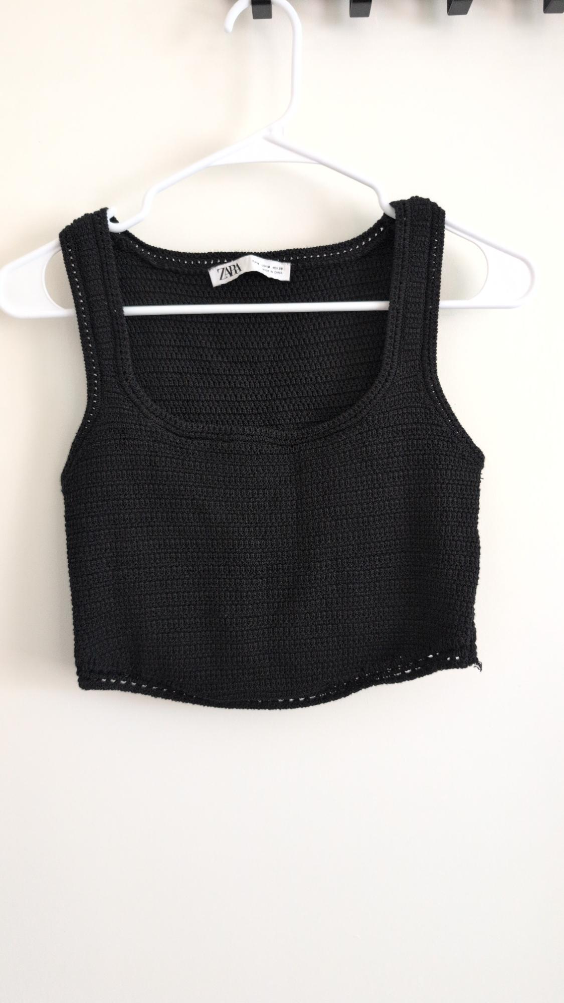 Black sleeveless knit cropped top hanging on a white hanger against a white wall. Label with the text ZARA.