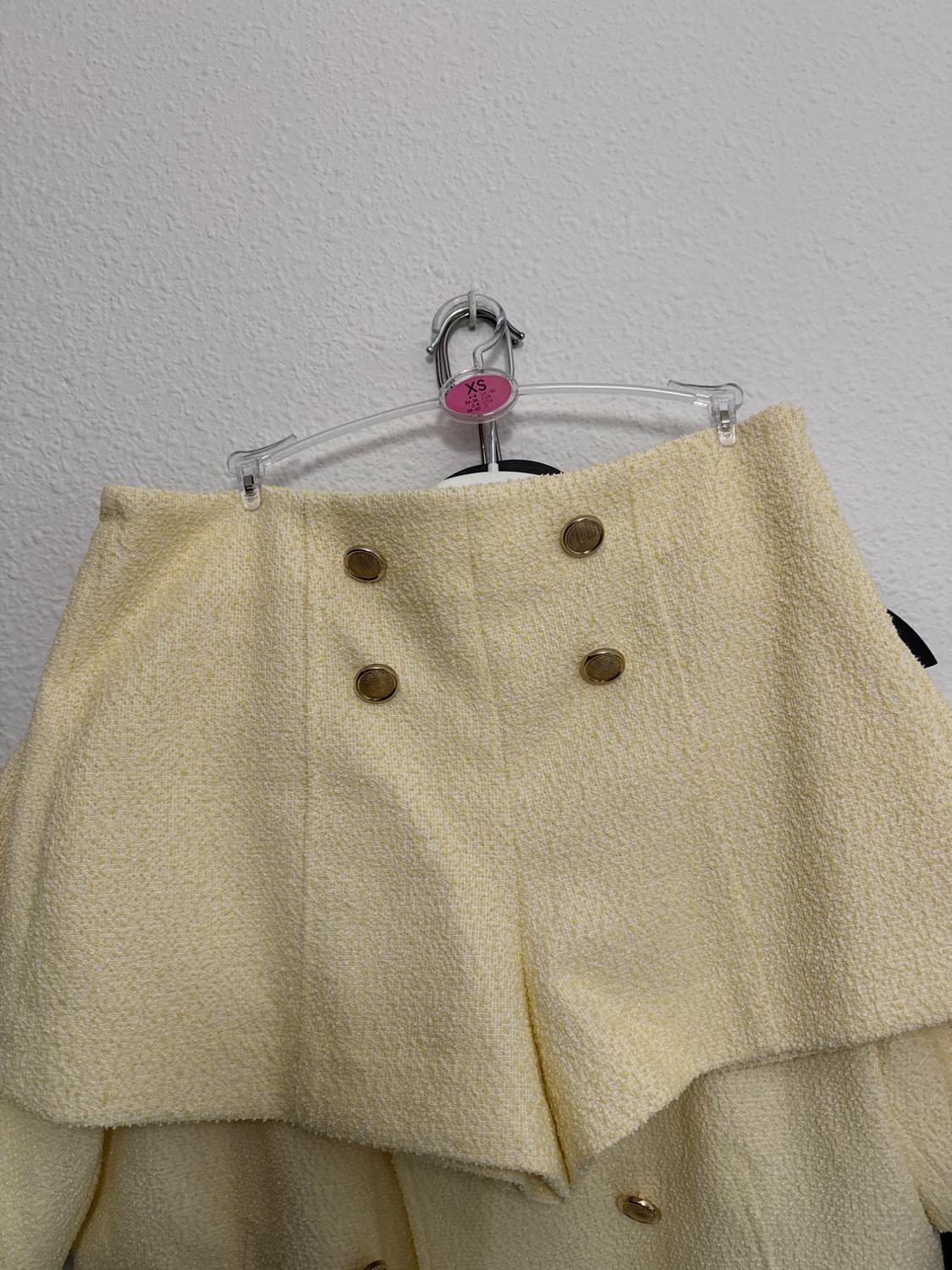 Yellow shorts with gold buttons, hanging on a clear hanger with a pink "XS" tag, against a white background.