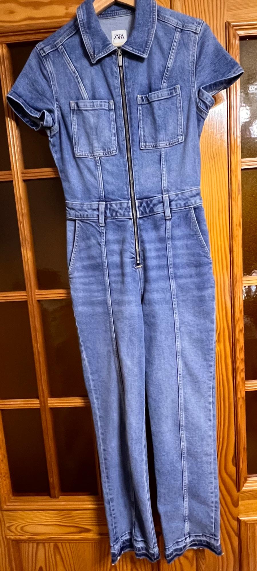 Blue denim jumpsuit with front zip and chest pockets, hanging on a wooden door. Visible label: ZARA.