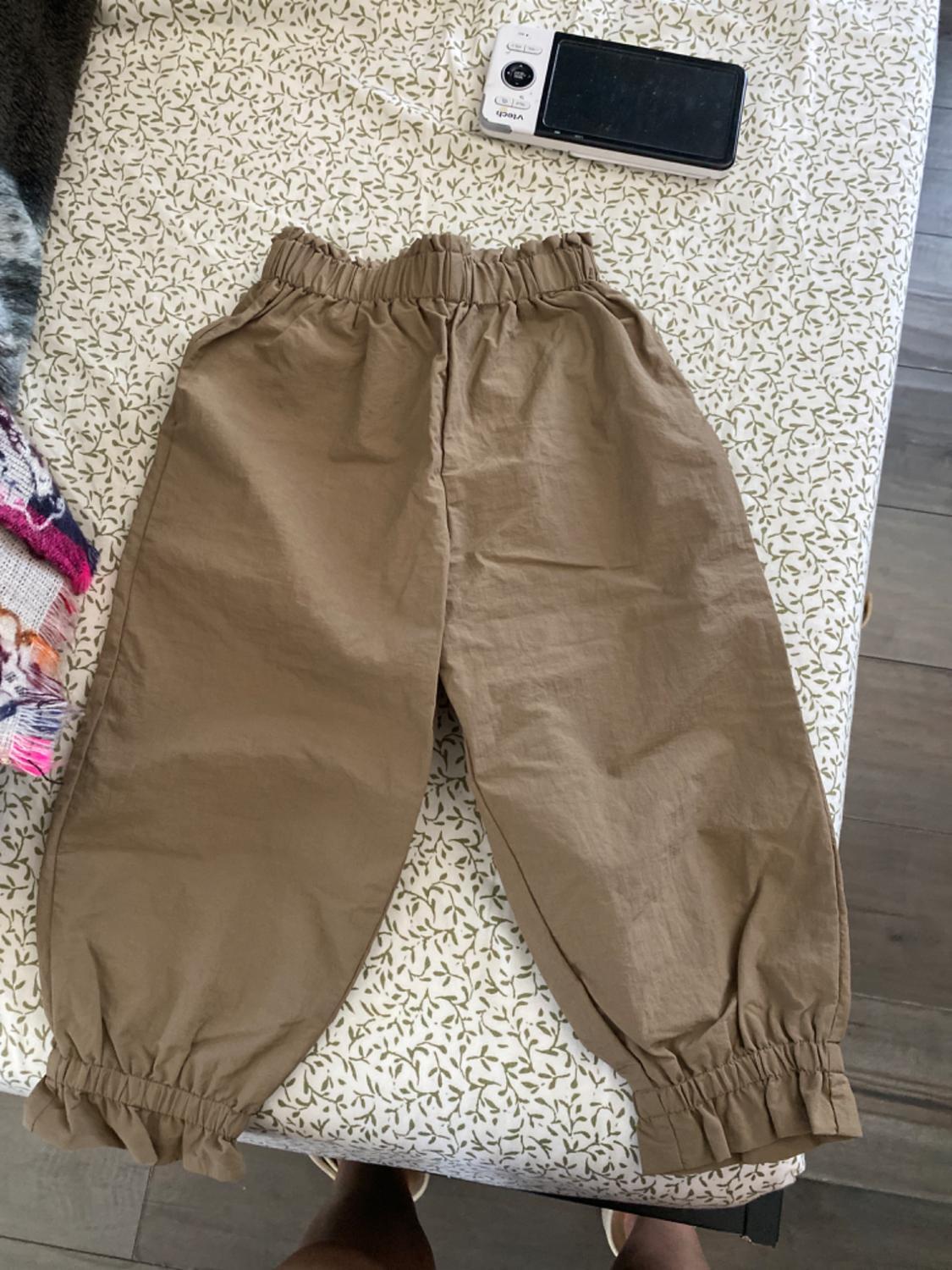 Light brown trousers with elasticated waist and ankles, on white fabric with a green leaf print. Black and white digital camera in the background.
