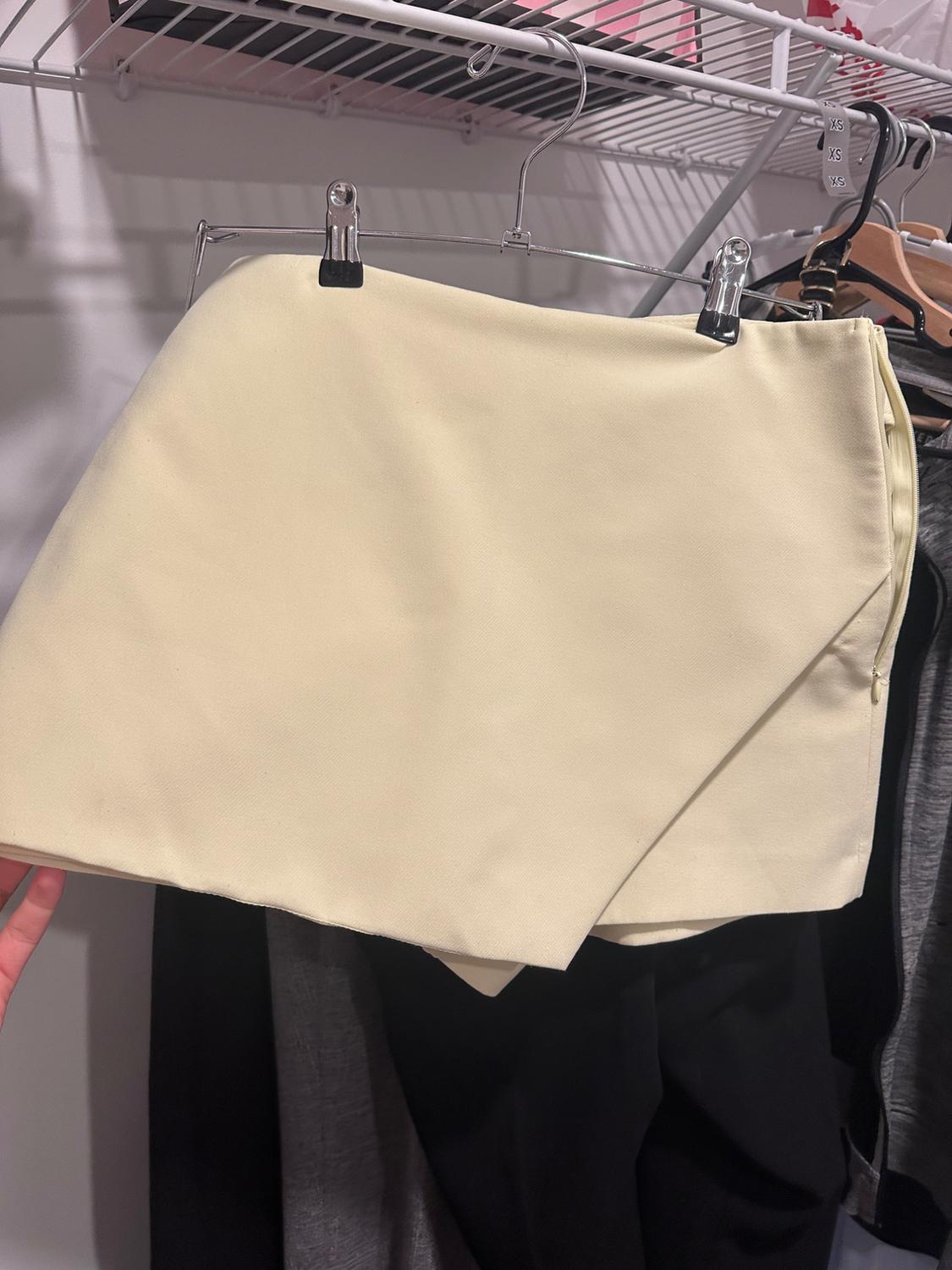 Cream asymmetric wrap skirt hanging on a hanger inside a wardrobe. The text XS XS XS is visible on the hanger. Casual style.