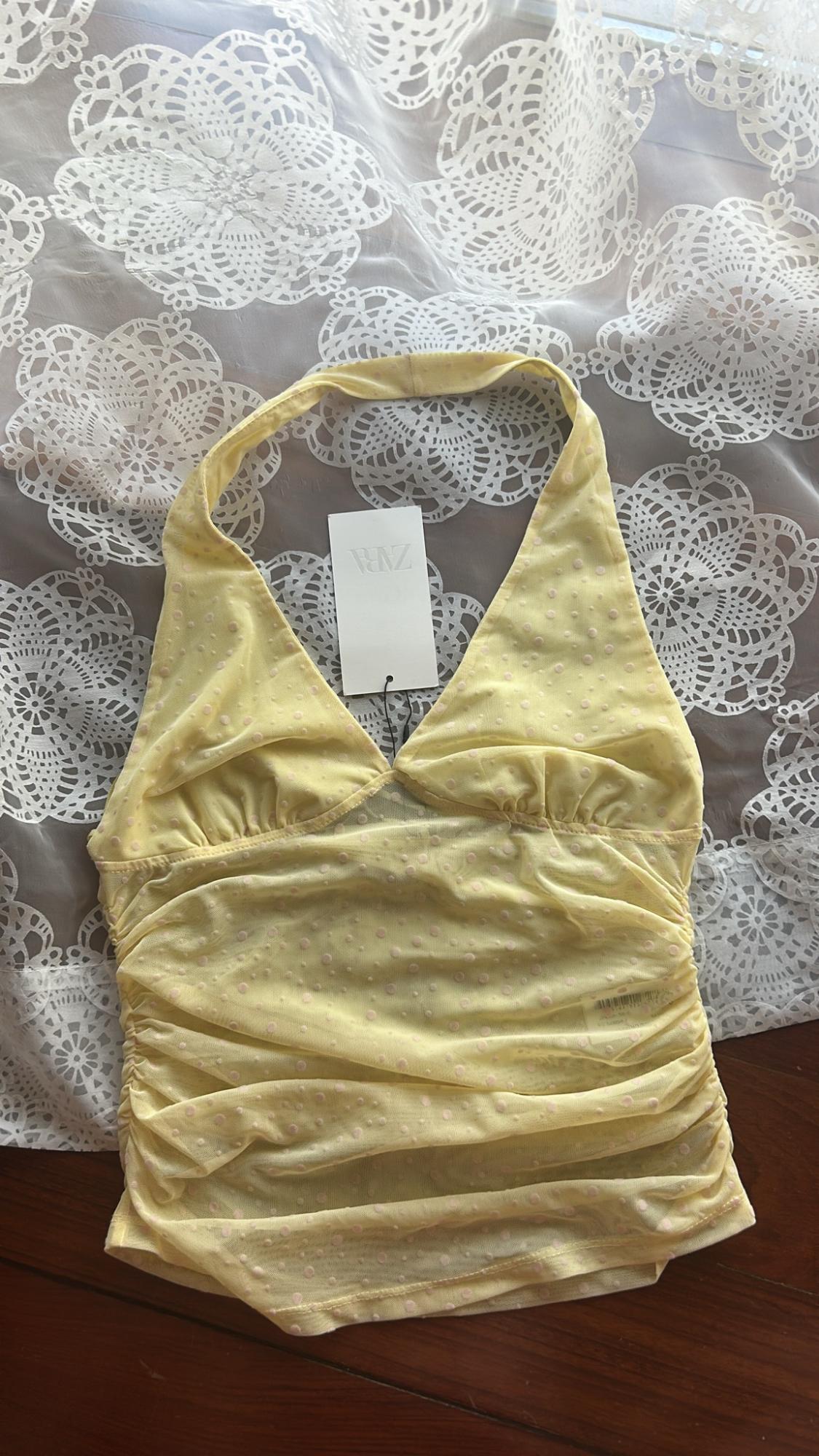 Yellow halterneck top with polka dots and ruching, on white lace fabric and a wooden surface. Label with ZARA text.