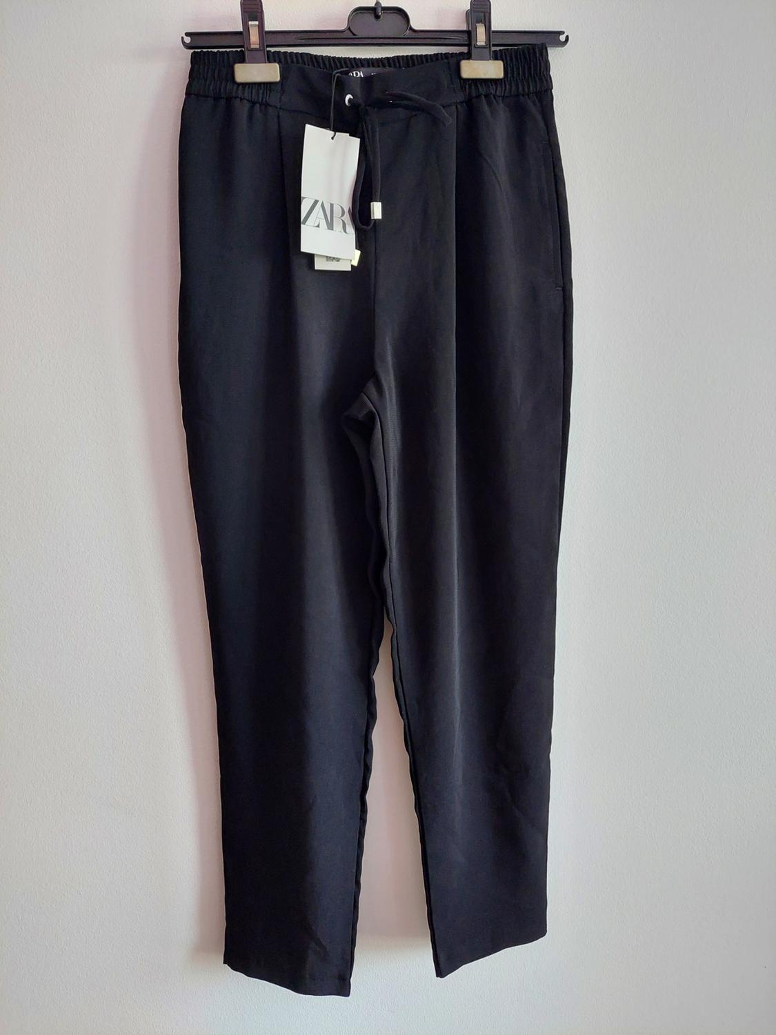 Black trousers with an elasticated waist and drawstring, hanging on a hanger against a light background. Includes a label with the text ZARA.