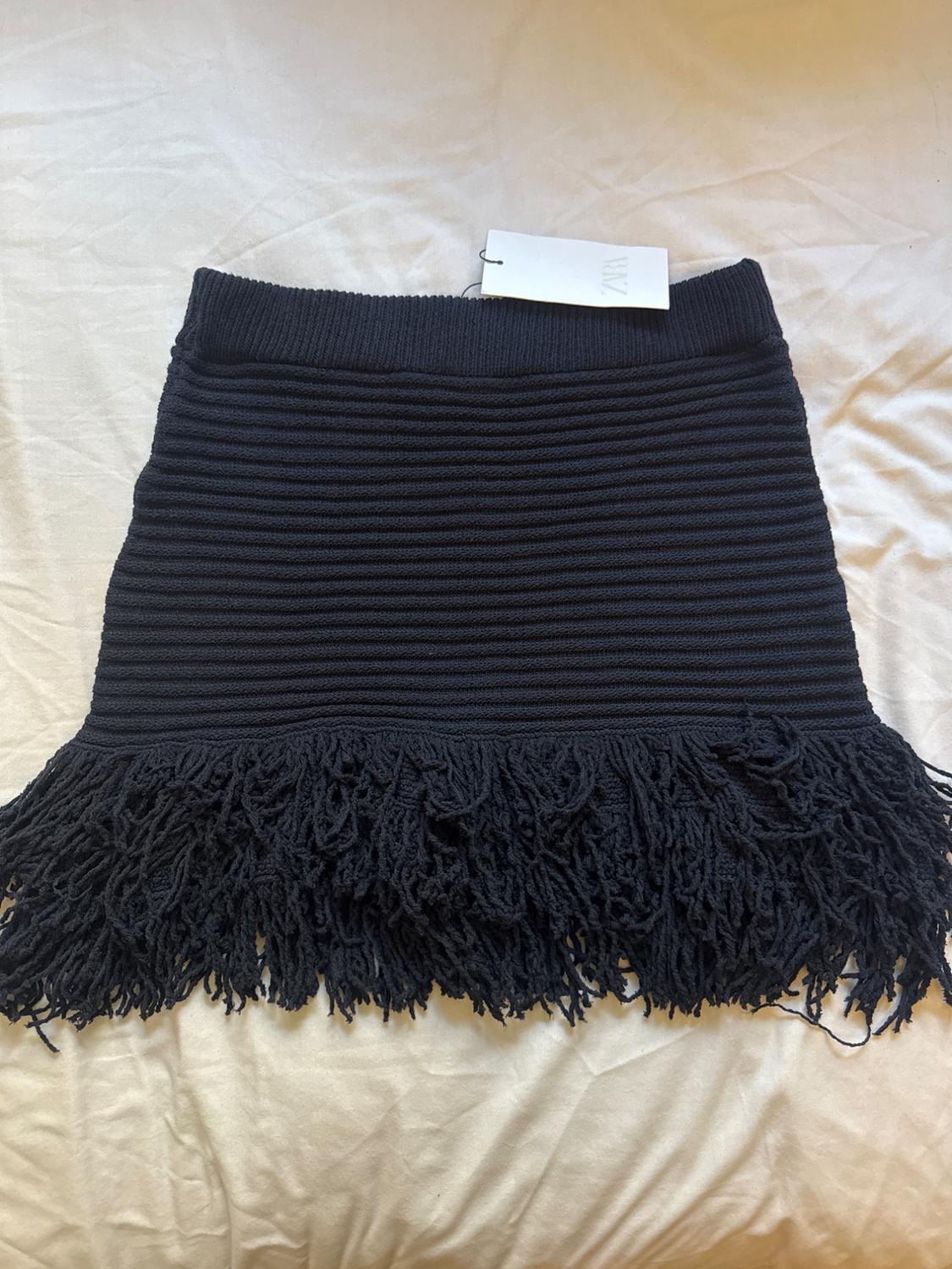 Black ribbed knit miniskirt with a chunky fringe hem, over a crumpled white fabric. Label with the text ZARA.