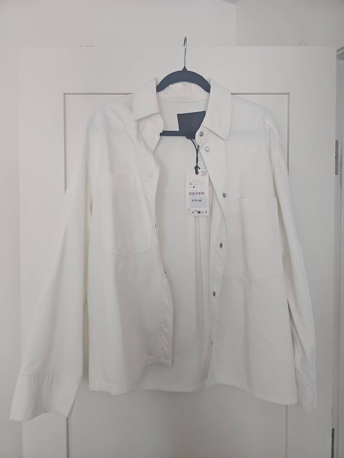 Long-sleeved white shirt with pockets, hanging on a white door. Label with the text S and $59.90.