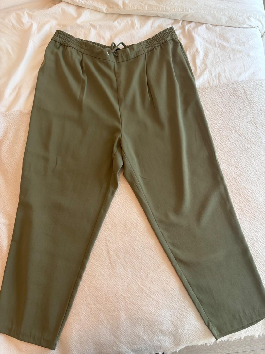 Olive green casual trousers with pleats, elasticated waist and drawstring, laid out on a bed with white sheets and a light textured blanket.