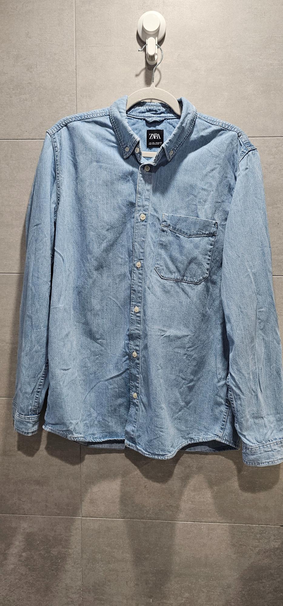 Blue long-sleeved denim shirt hanging on grey tiles. Text: ZARA EUR L USA L MEX 42 MADE IN BANGLADESH.