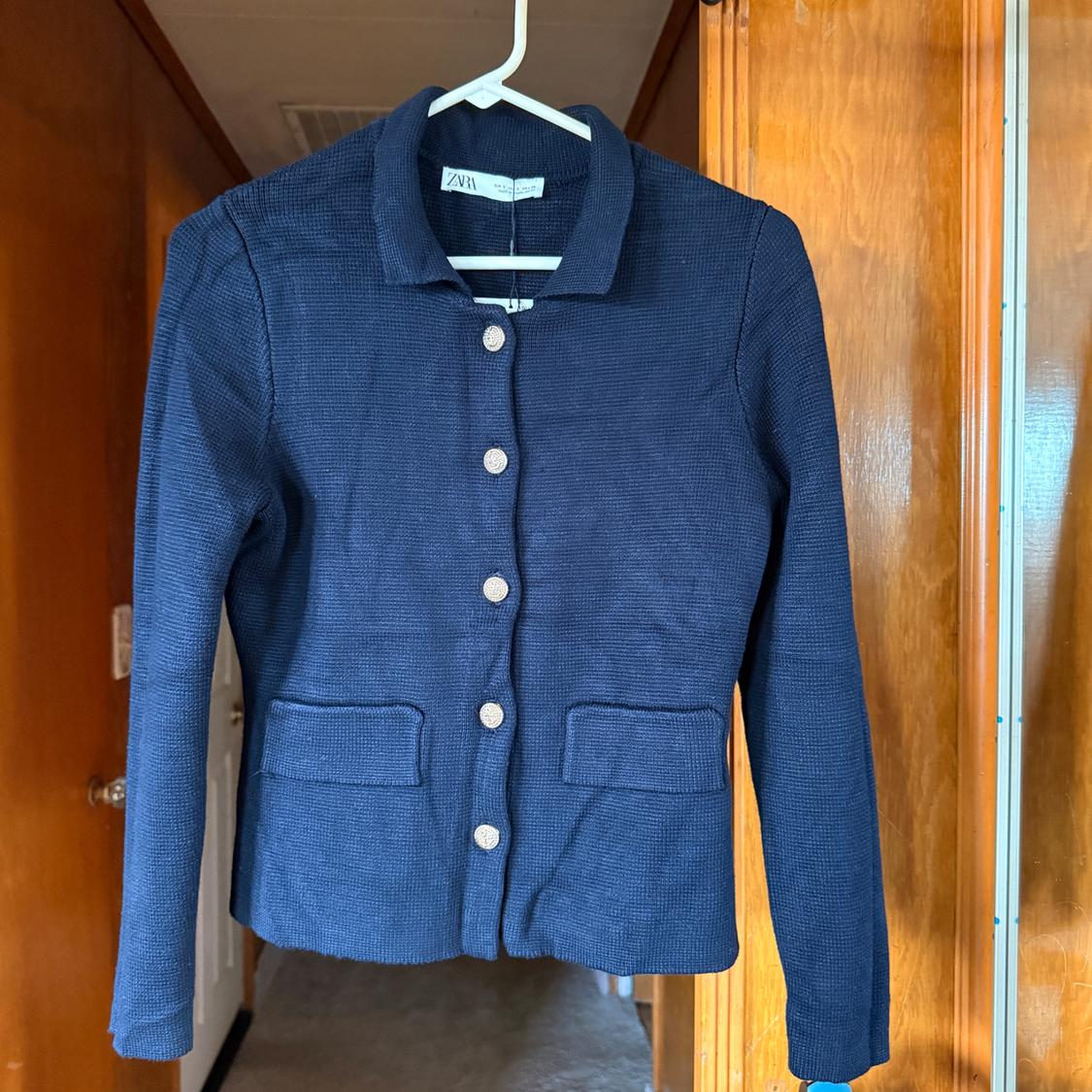 Blue jacket hanging on a white hanger, with silver buttons and two front pockets. Visible tag with the "Zara" brand.