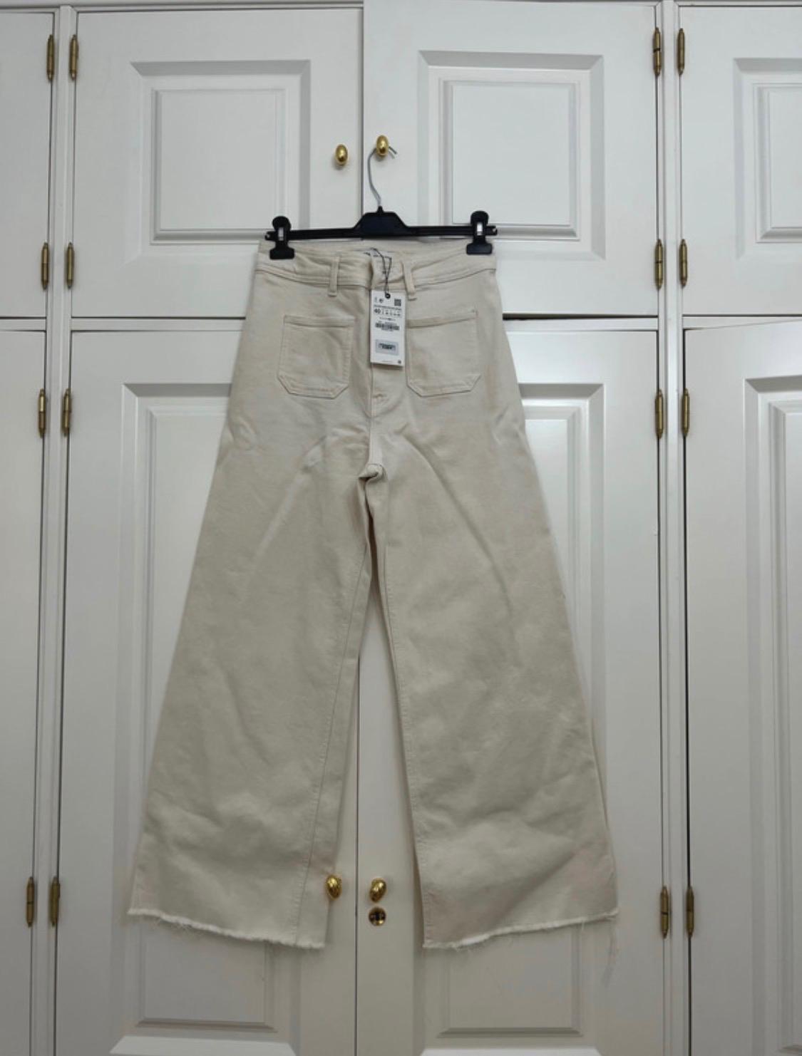 Cream-coloured wide-leg trousers with front pockets and a frayed hem, hanging on a black hanger in front of a white wardrobe.