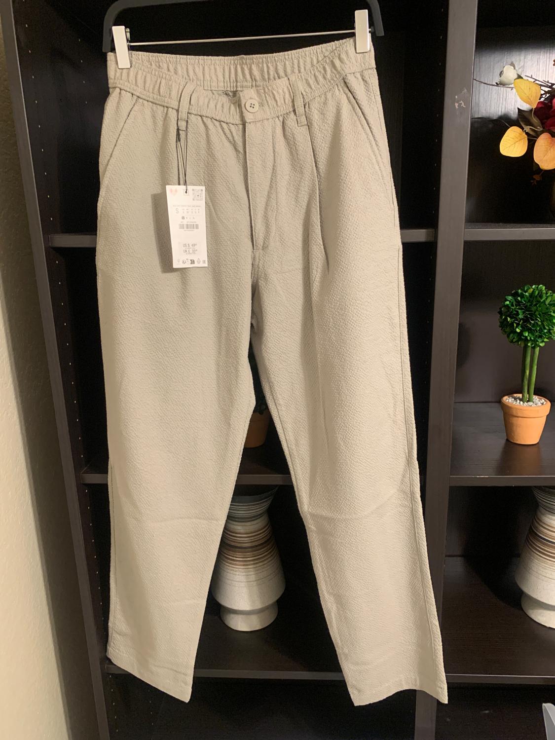 Beige trousers hanging on a clothes rail, with a visible label. Dark shelving unit background with decorative plant pots.