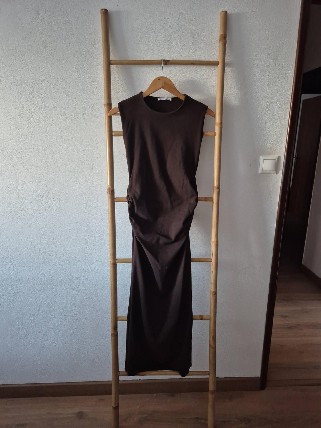 Long brown sleeveless dress with side ruching, hanging on a bamboo ladder leaning against a white wall.